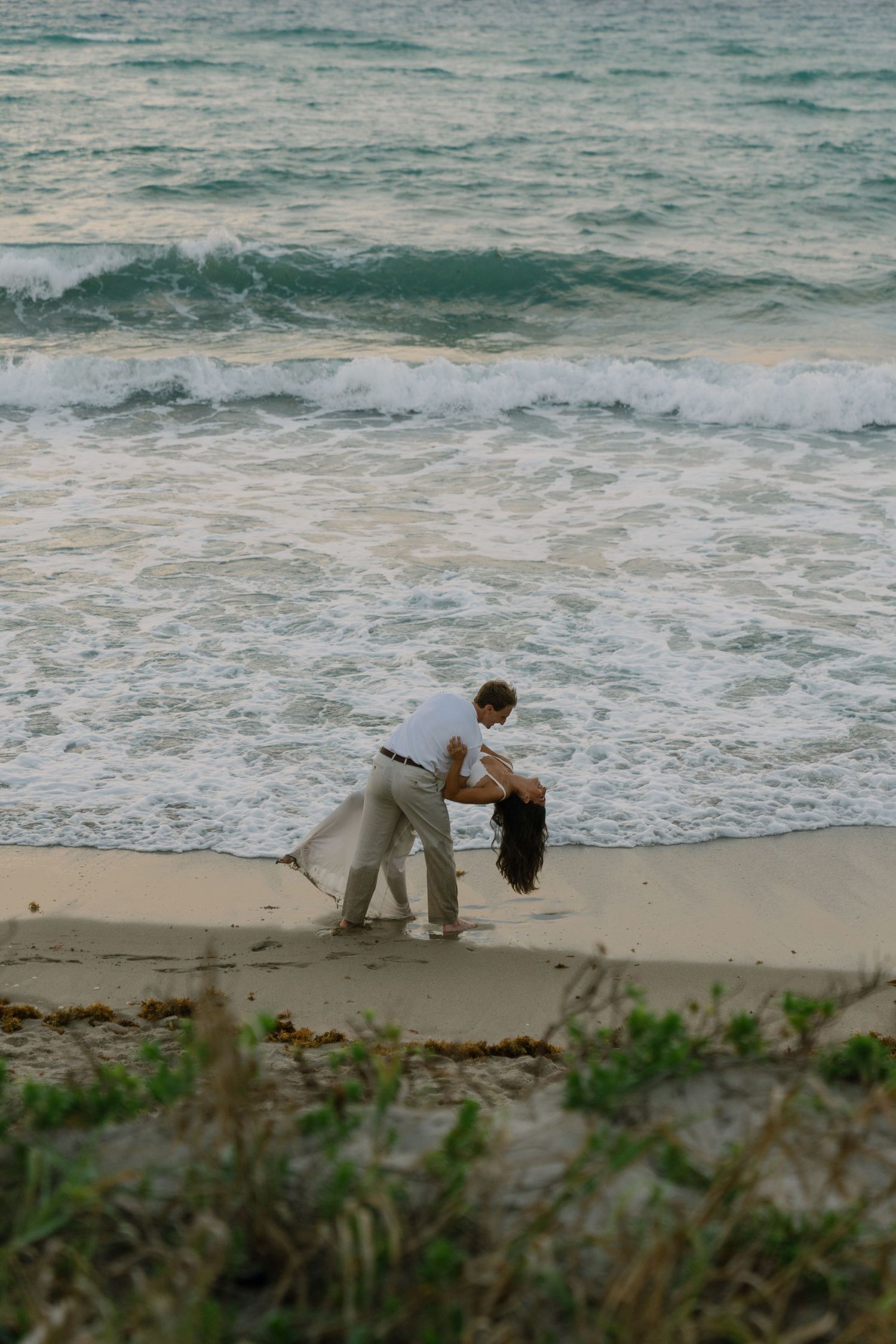 Couple dancing romantically on a sandy beach at dusk with ocean waves crashing behind them.