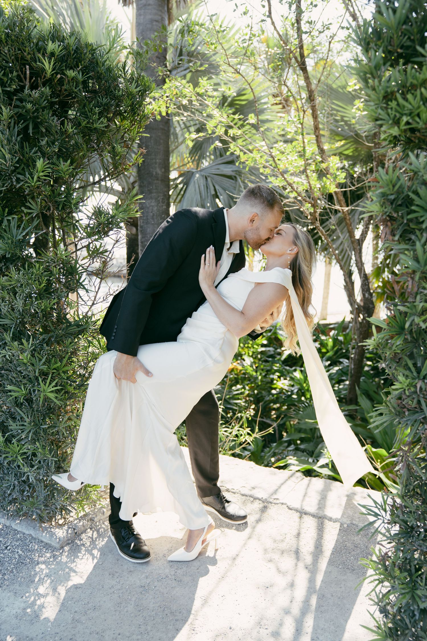 Romantic couple in wedding attire sharing passionate dips and kisses in a lush tropical garden setting.