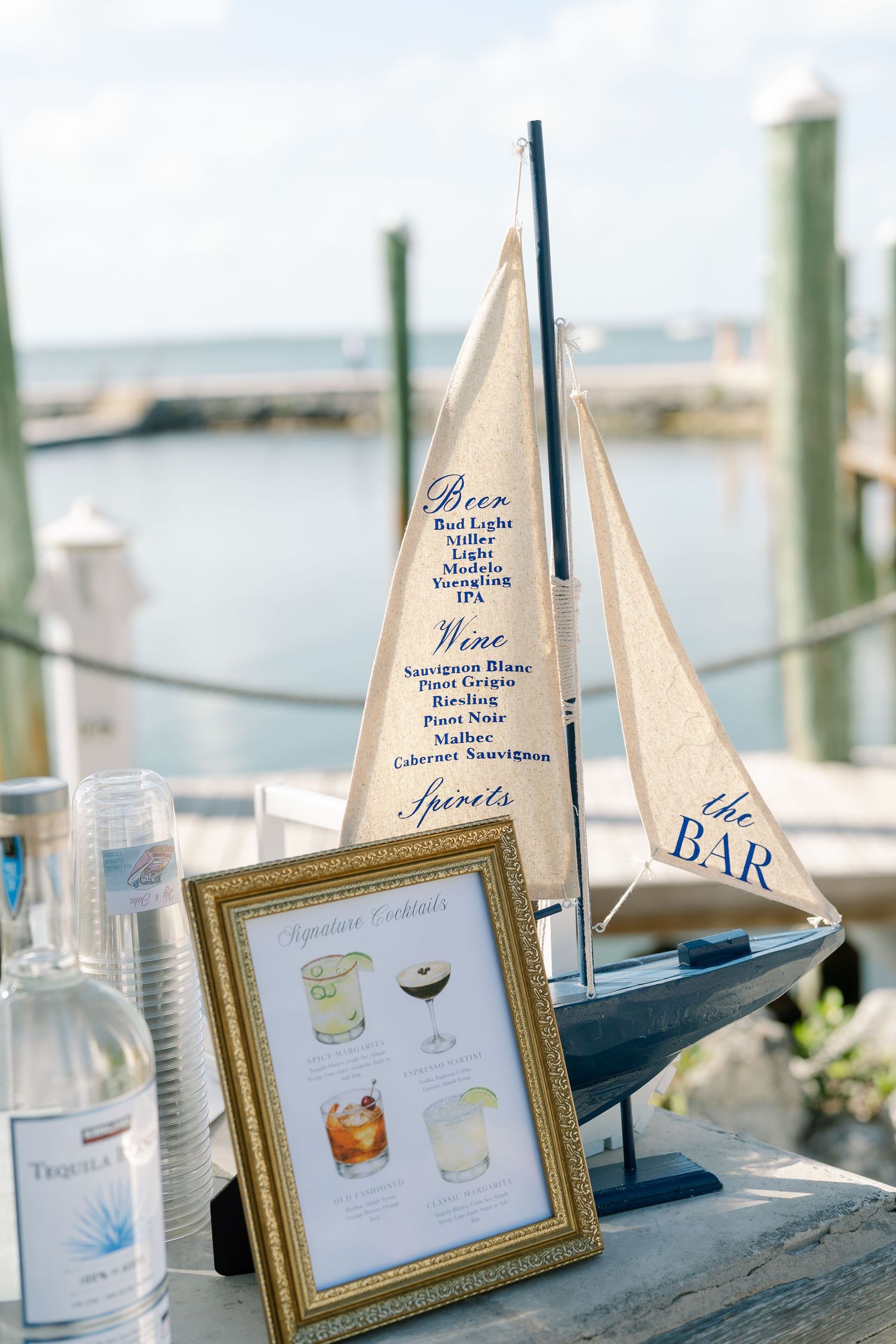 A decorative sailboat model and a framed cocktail menu displayed on a table, with a coastal backdrop.