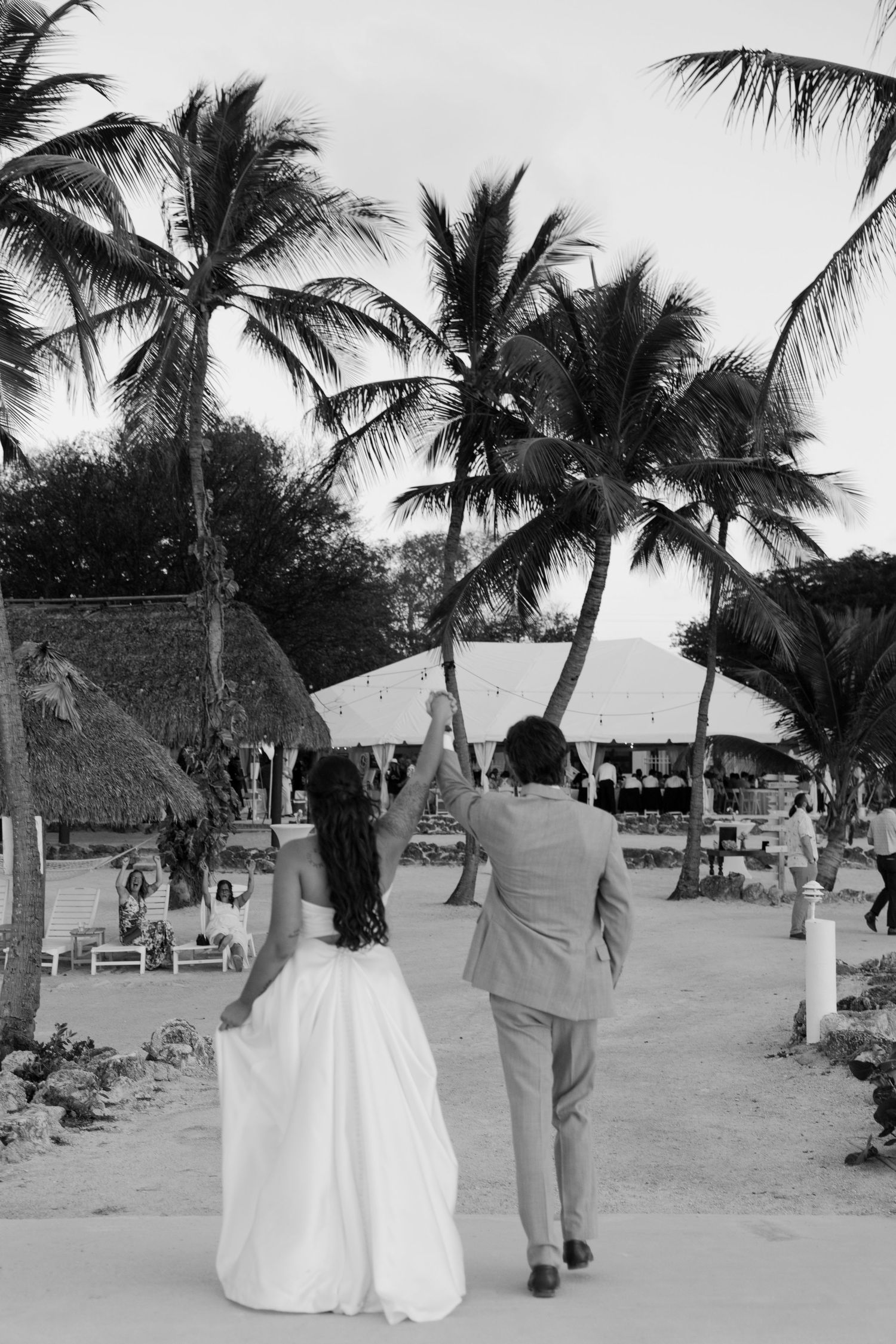 A black and white image of a bride and groom walking on a beach with palm trees.