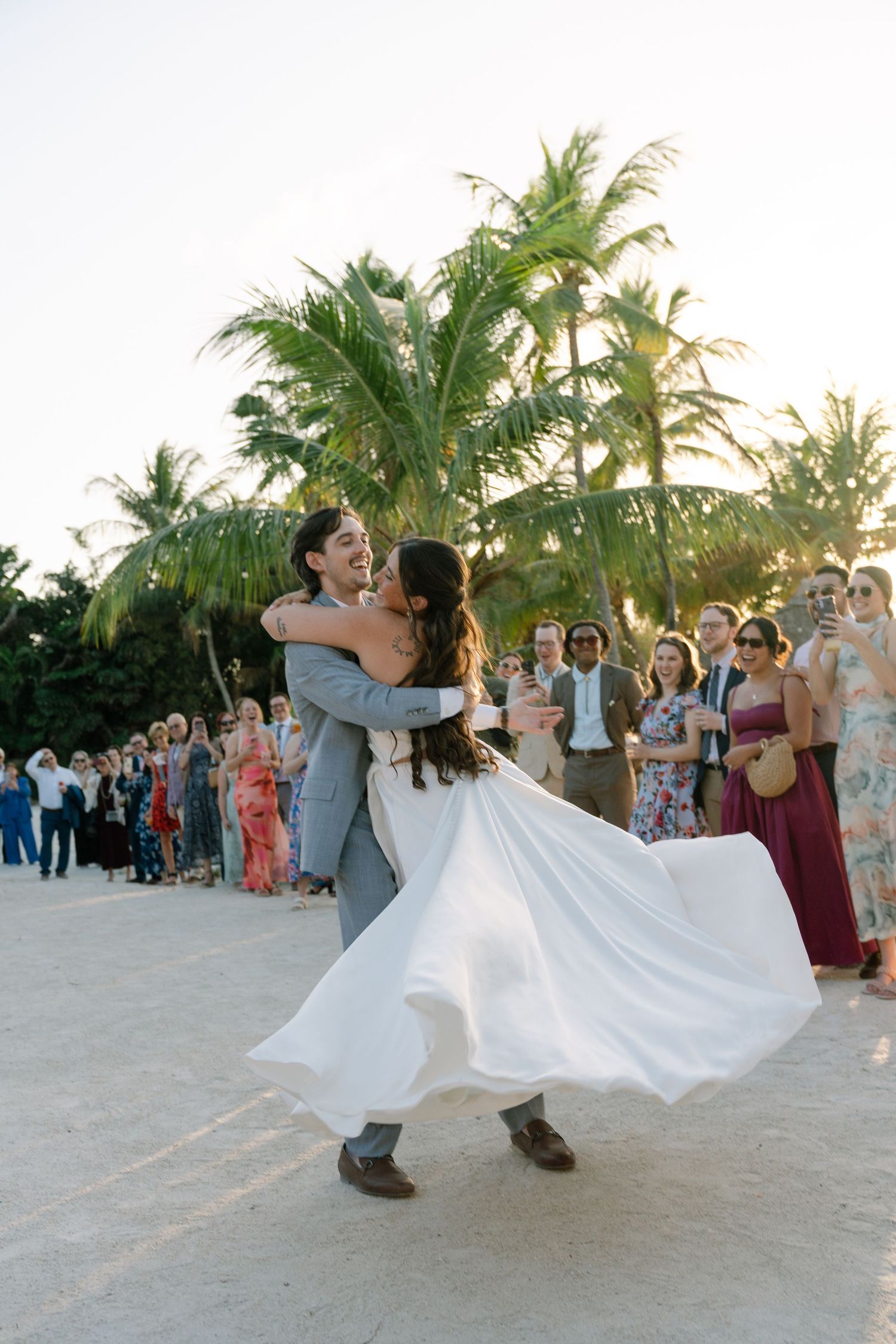 Groom lifts bride in white gown on sandy beach, guests watching, palm trees at sunset during outdoor wedding celebration.