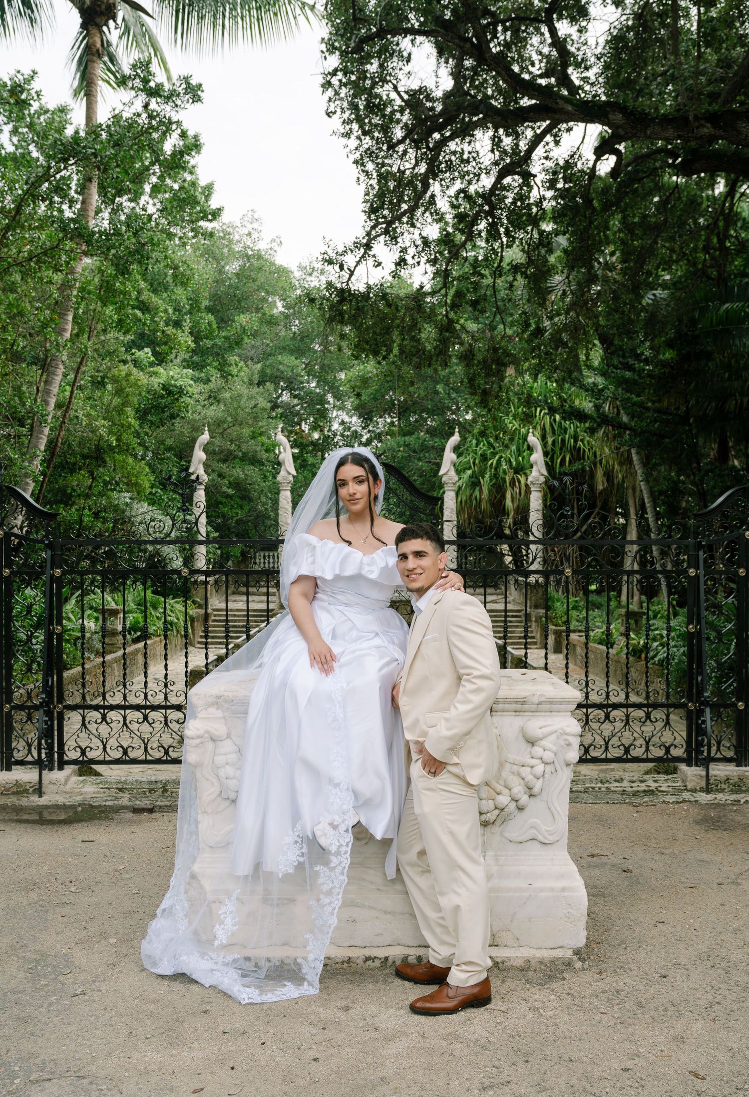 Elegant wedding couple poses by ornate iron gates in a lush tropical garden setting with palm trees and greenery.