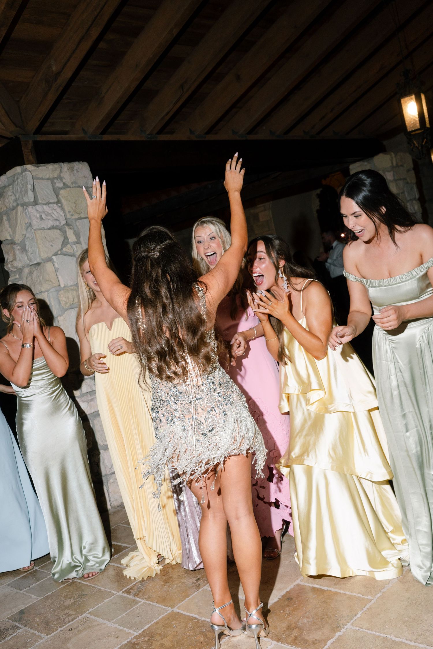 Women in elegant gowns dancing and celebrating at a wedding reception on a stone patio.