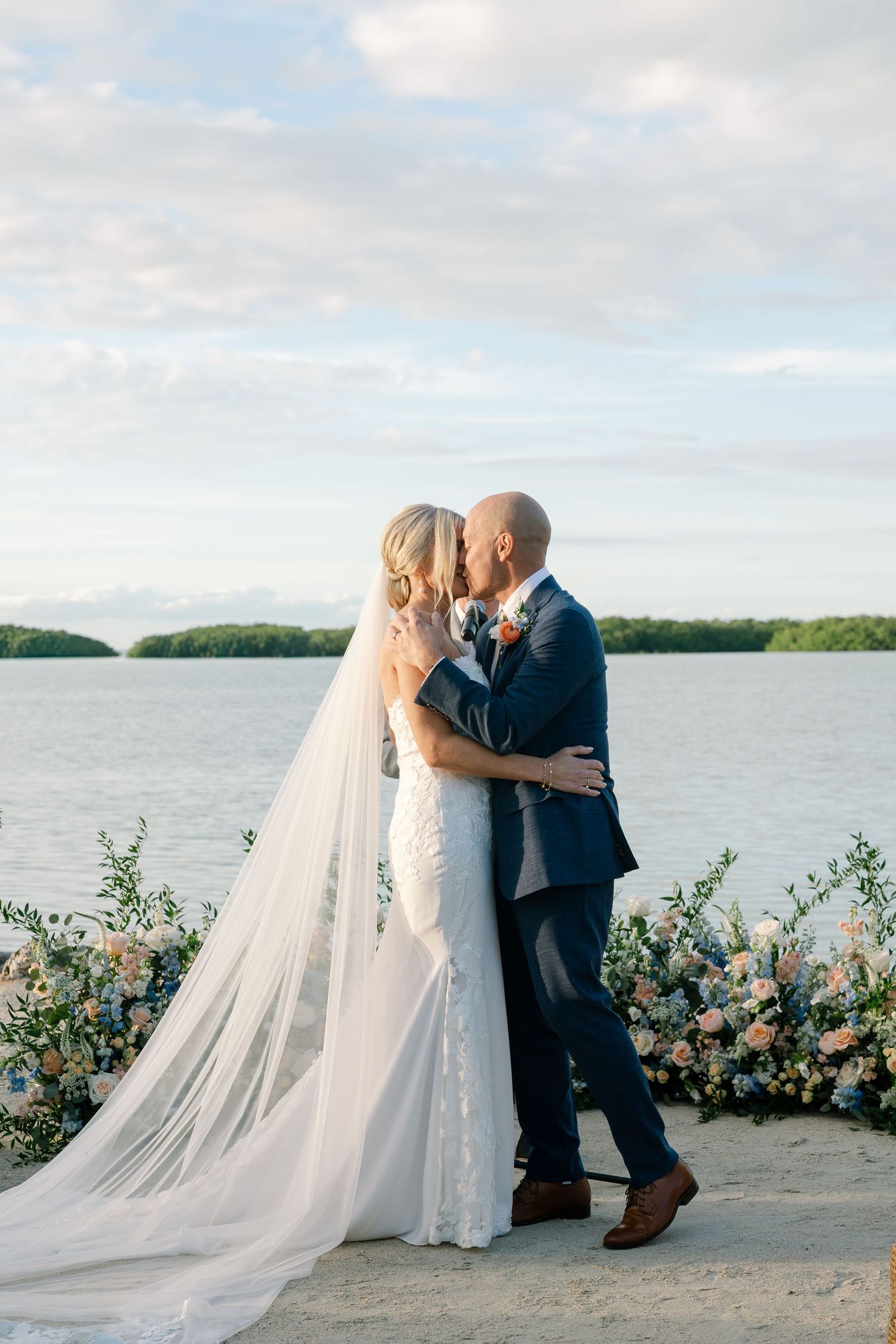 A romantic wedding portrait of a couple embracing by the water with floral decorations and cloudy skies.