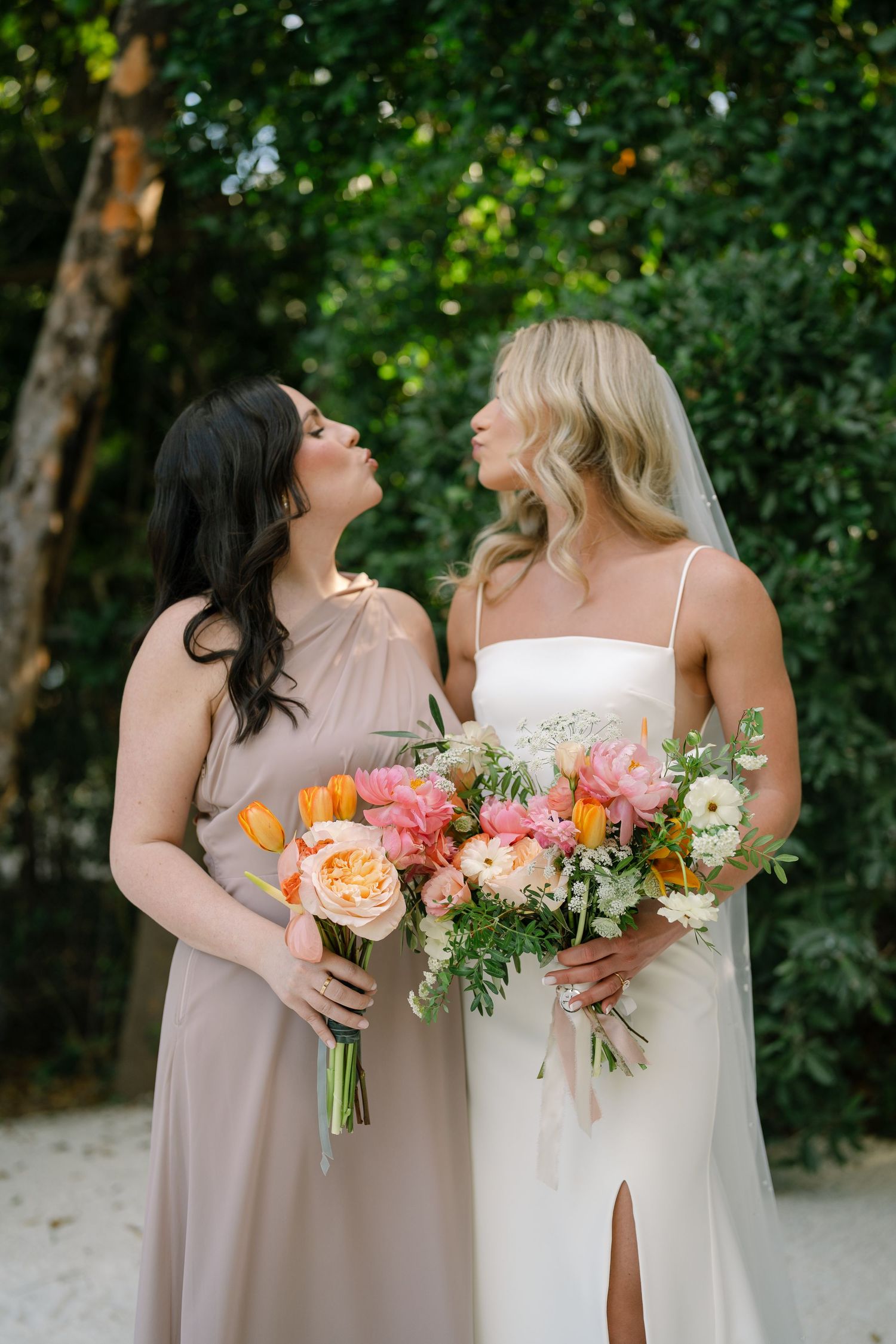 Two attendants in neutral dresses sharing a moment while holding colorful bouquets outdoors.