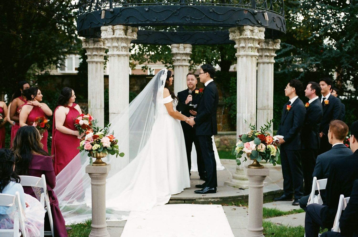 A romantic outdoor wedding ceremony takes place in a garden setting with stone columns and floral arrangements.