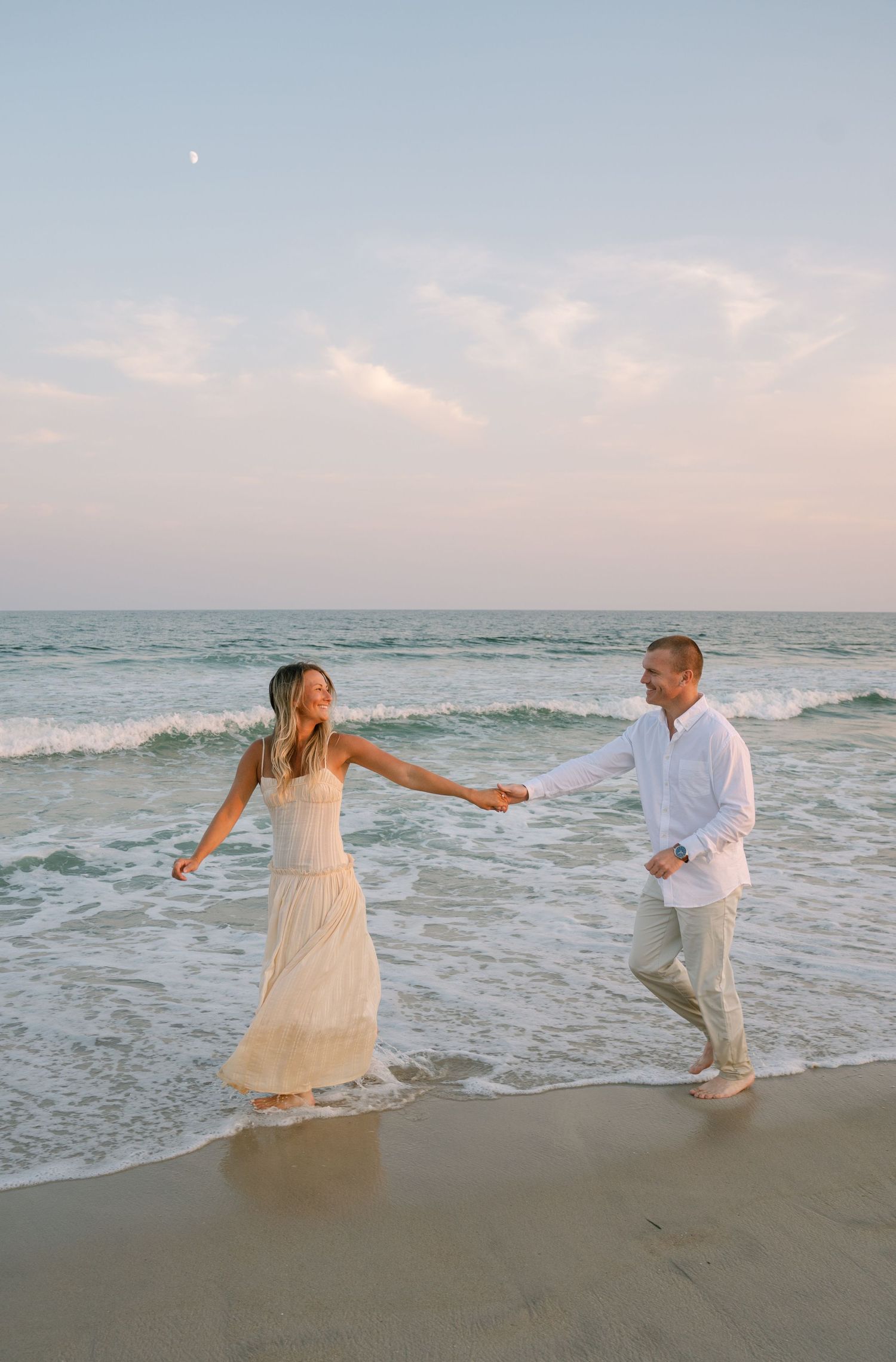 A couple in white attire dances playfully along the ocean shore at sunset.