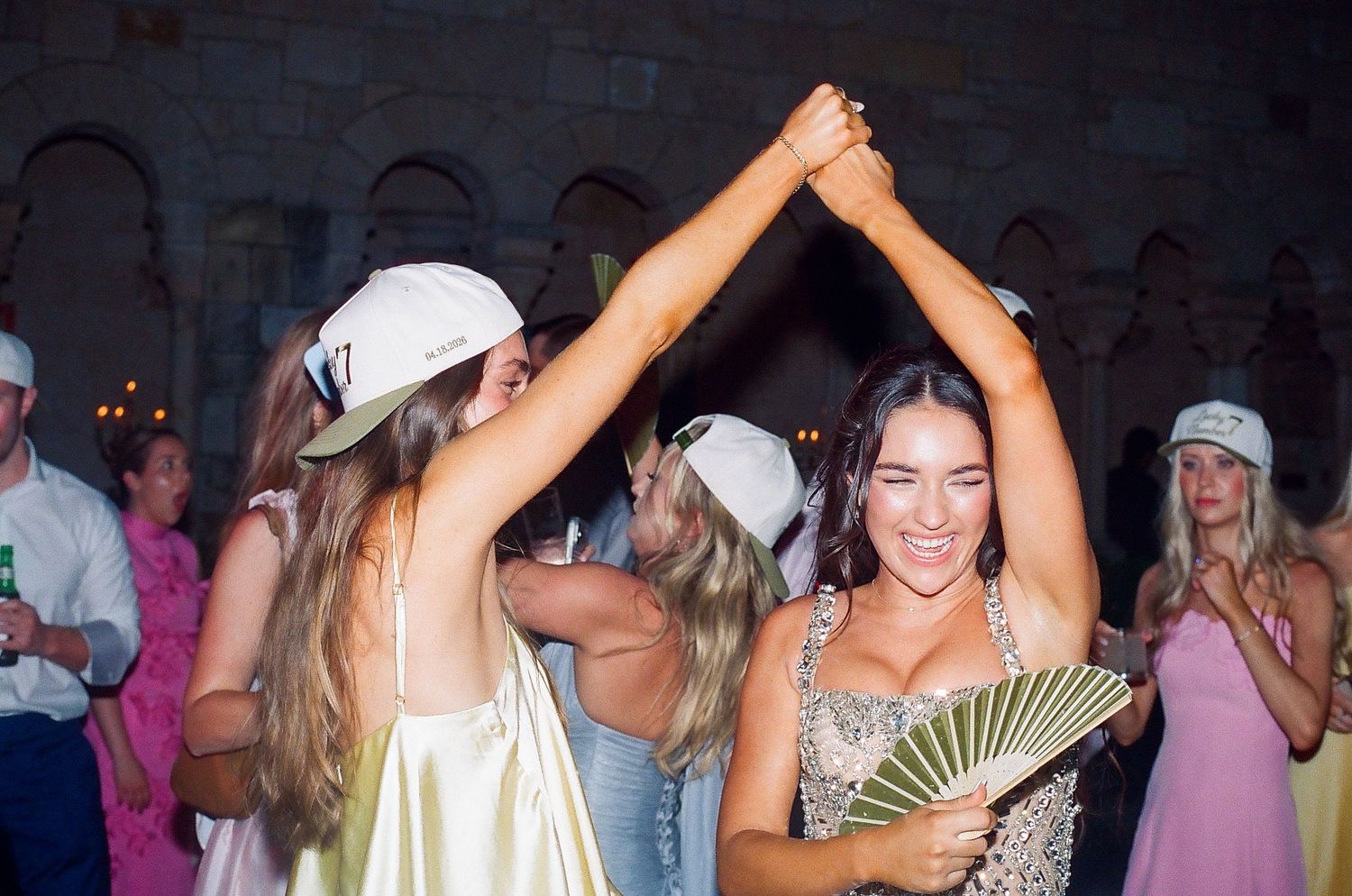 Bride dancing joyfully with friend at wedding reception, holding green fan, wearing sparkly dress indoors.
