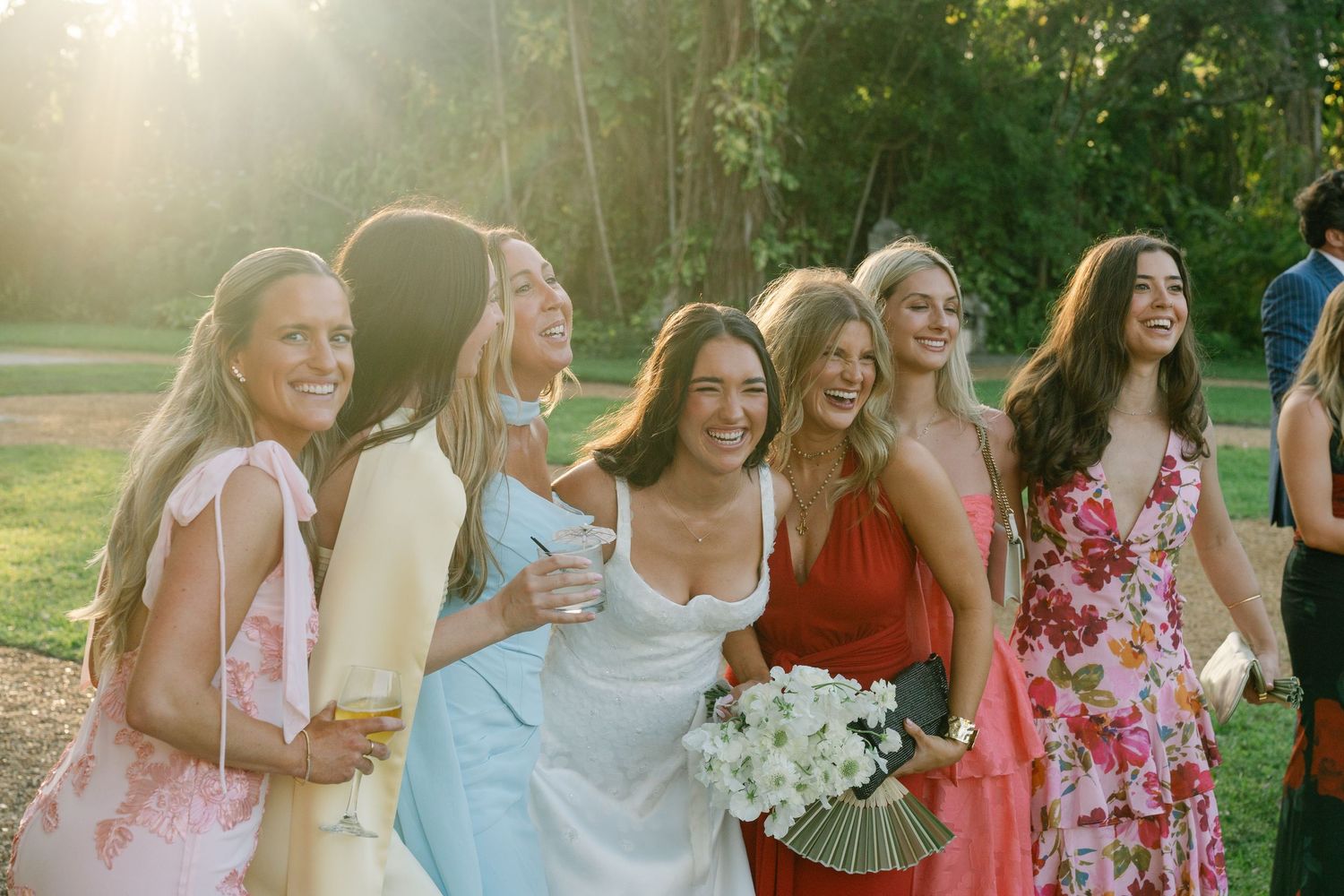 Bride and bridesmaids laughing together outdoors in colorful dresses during golden hour wedding celebration.