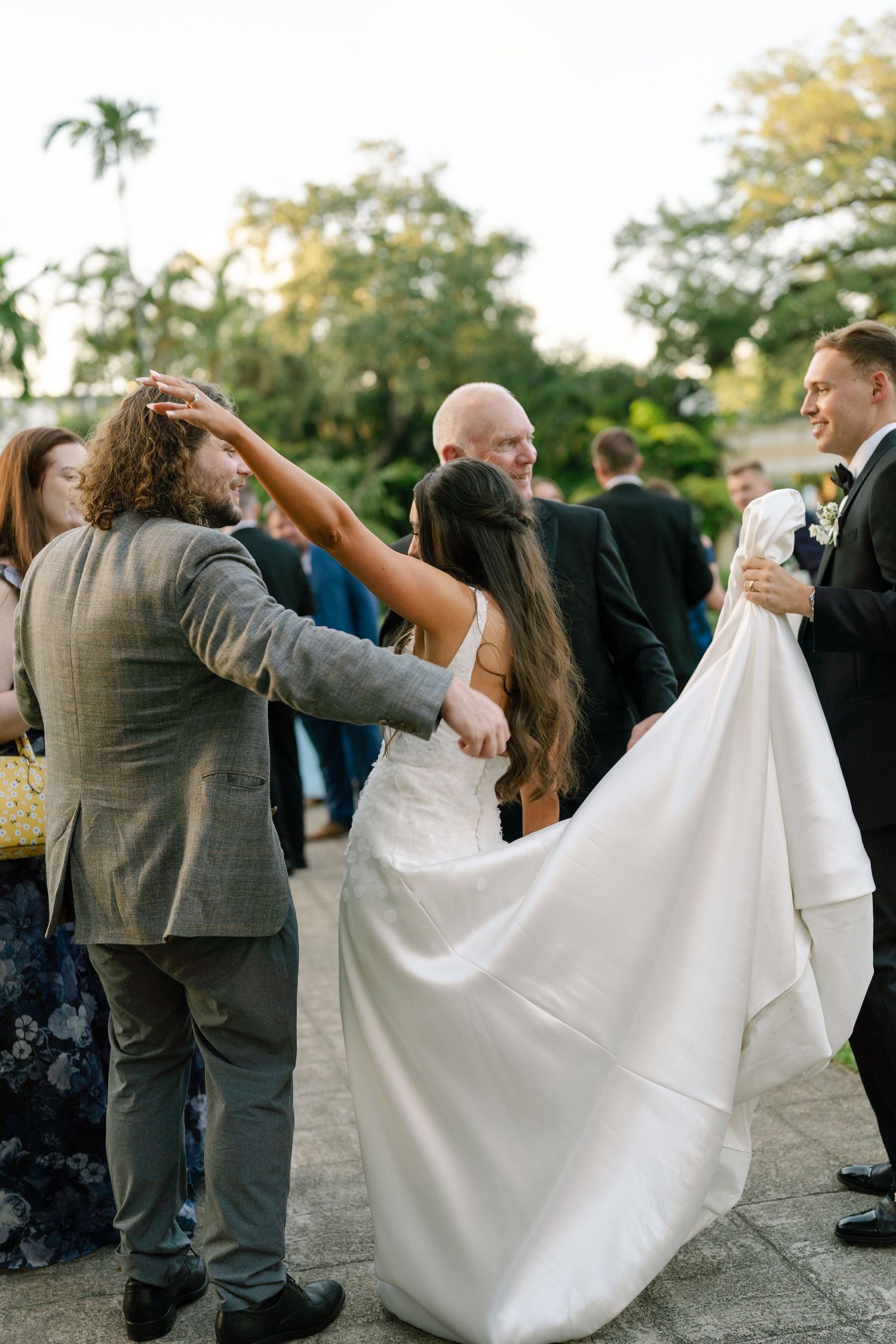 Wedding guests celebrate outdoors as bride dances with guest while groom watches and smiles at elegant garden reception.