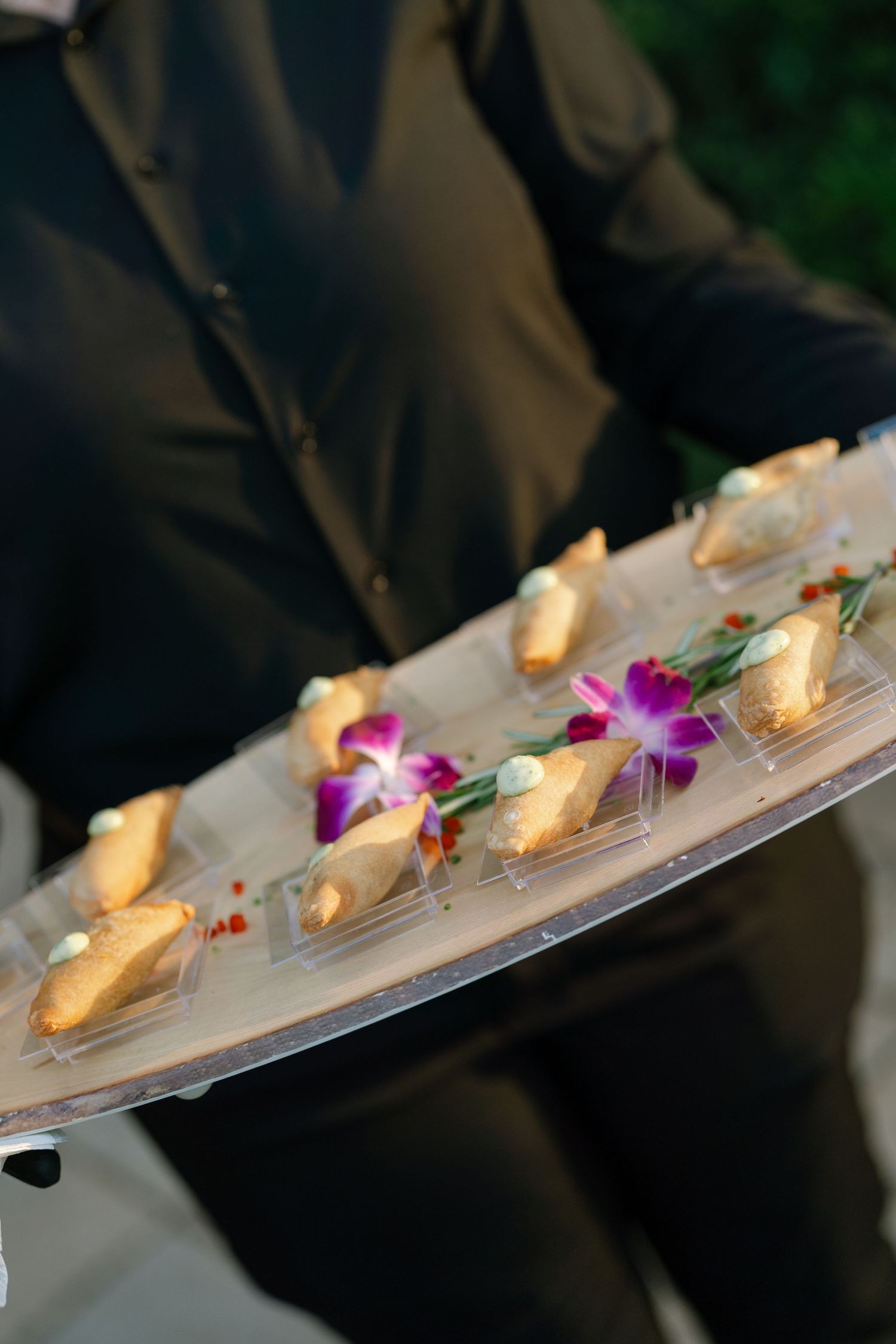 Server in black uniform presenting elegant tray of dessert canapes garnished with purple orchid flowers.