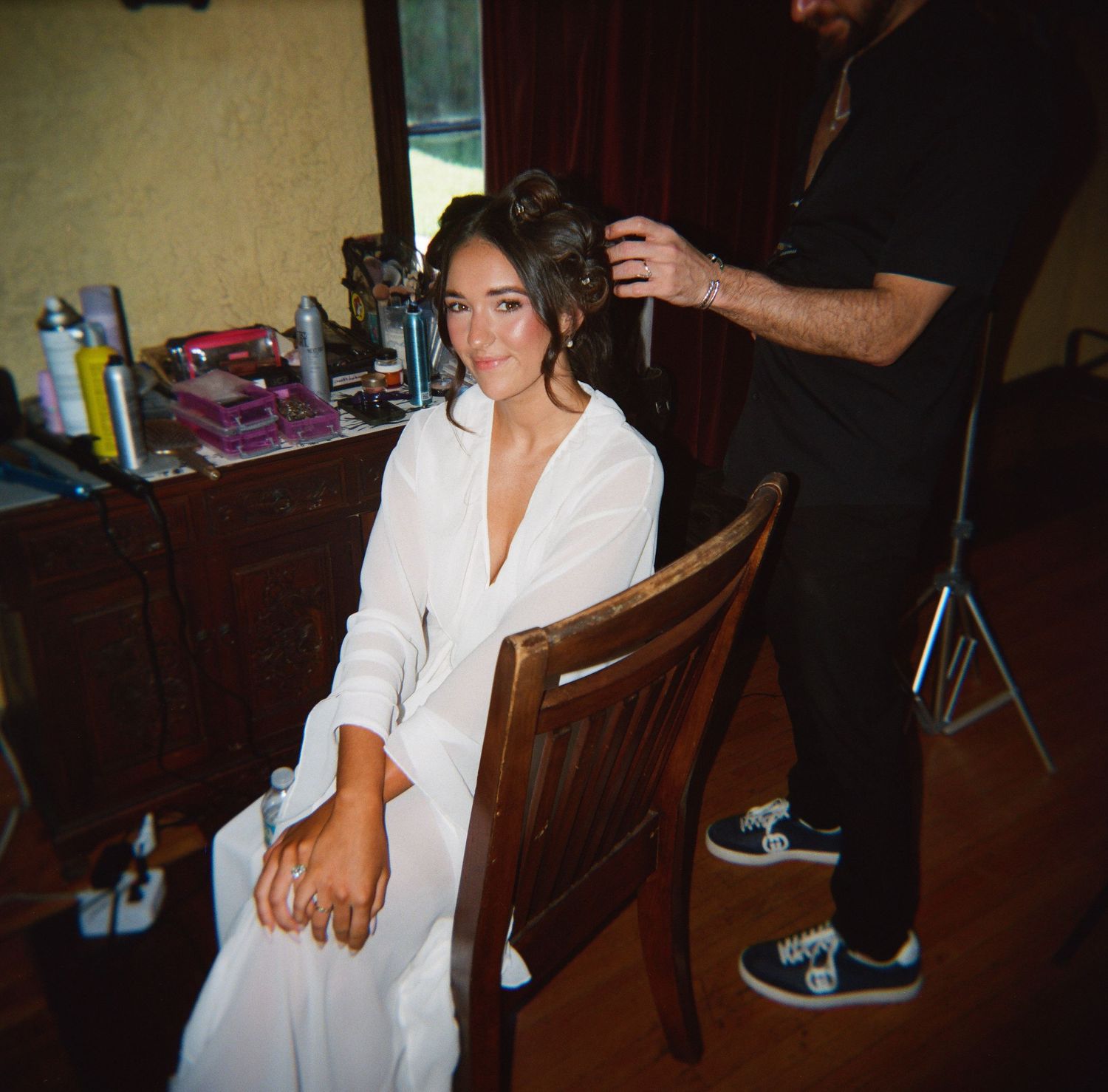 Bride in white robe sits in wooden chair while hairstylist works on her updo in a vintage dressing room.
