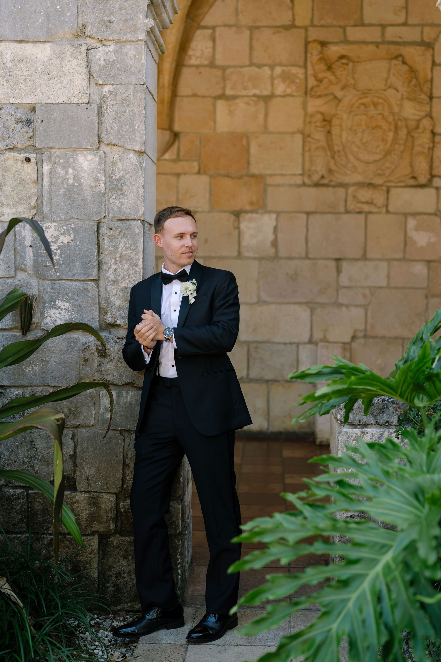 Groom in black tuxedo adjusts cufflinks while leaning against ornate stone wall with tropical plants.