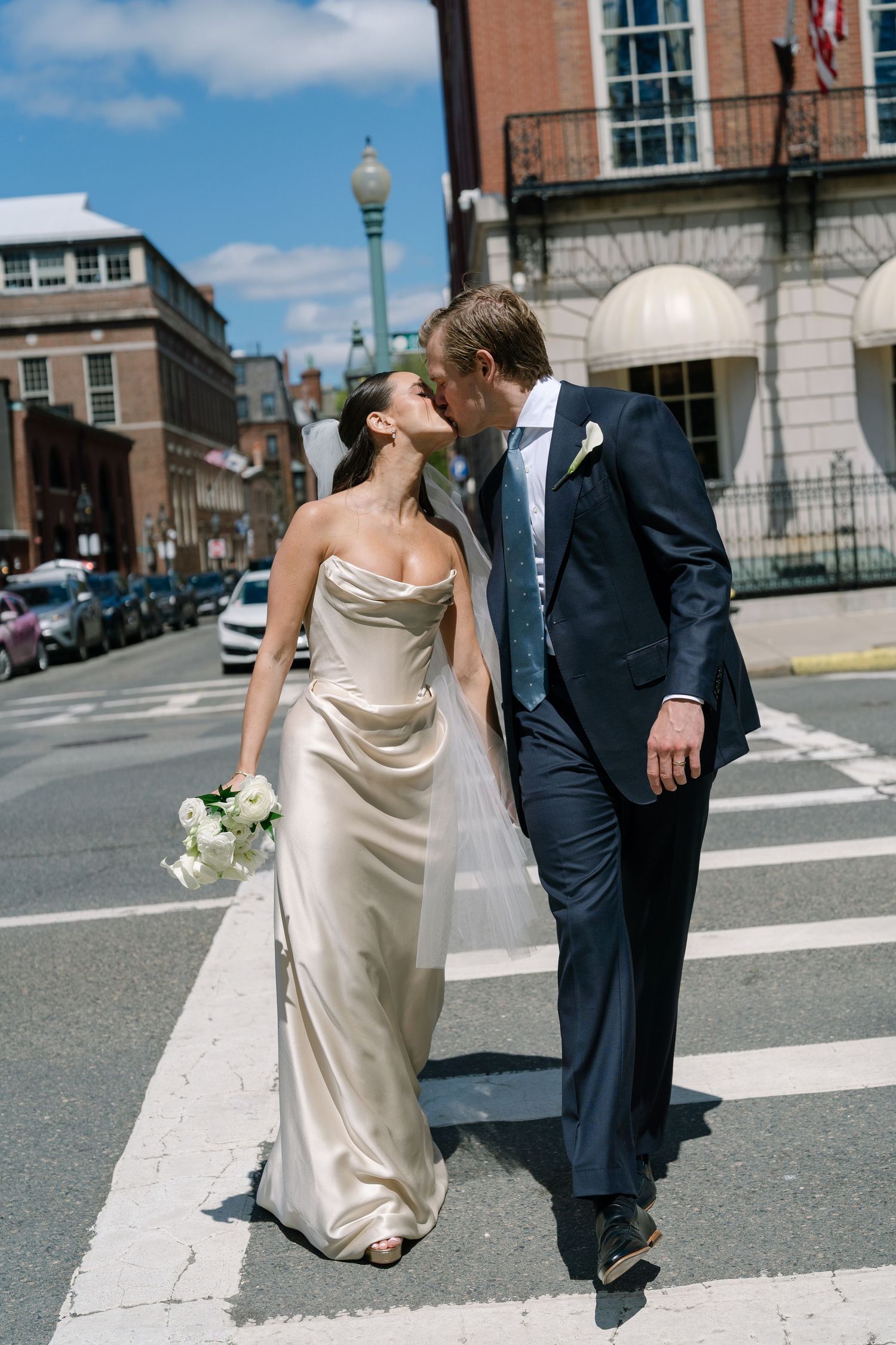 Bride and groom share a kiss while crossing a sunny city street, she in a champagne satin gown holding white flowers.
