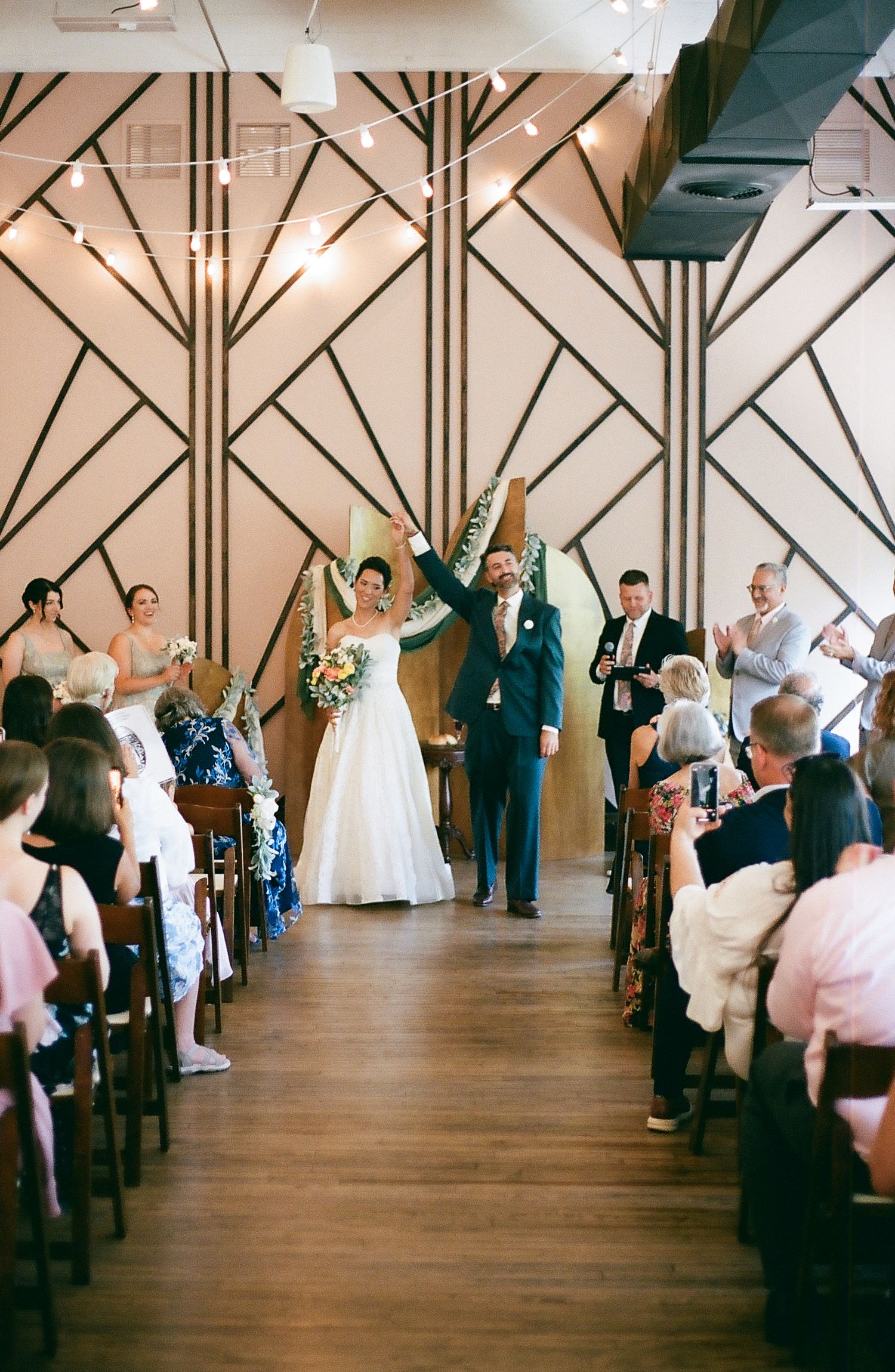 Wedding ceremony taking place in a modern venue with geometric wall patterns and wooden floors.