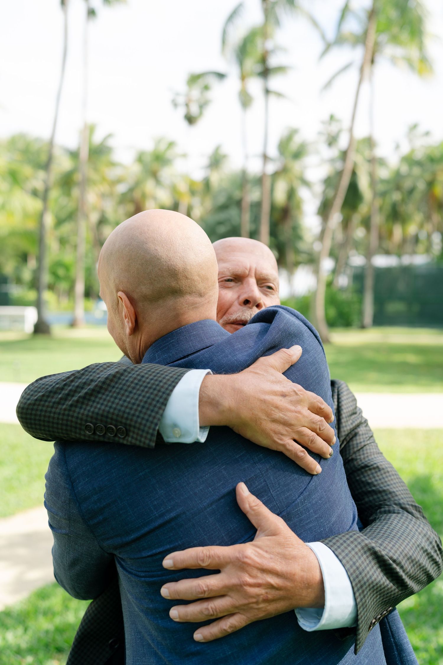 Two men in suits share an emotional embrace outdoors with palm trees in the background.