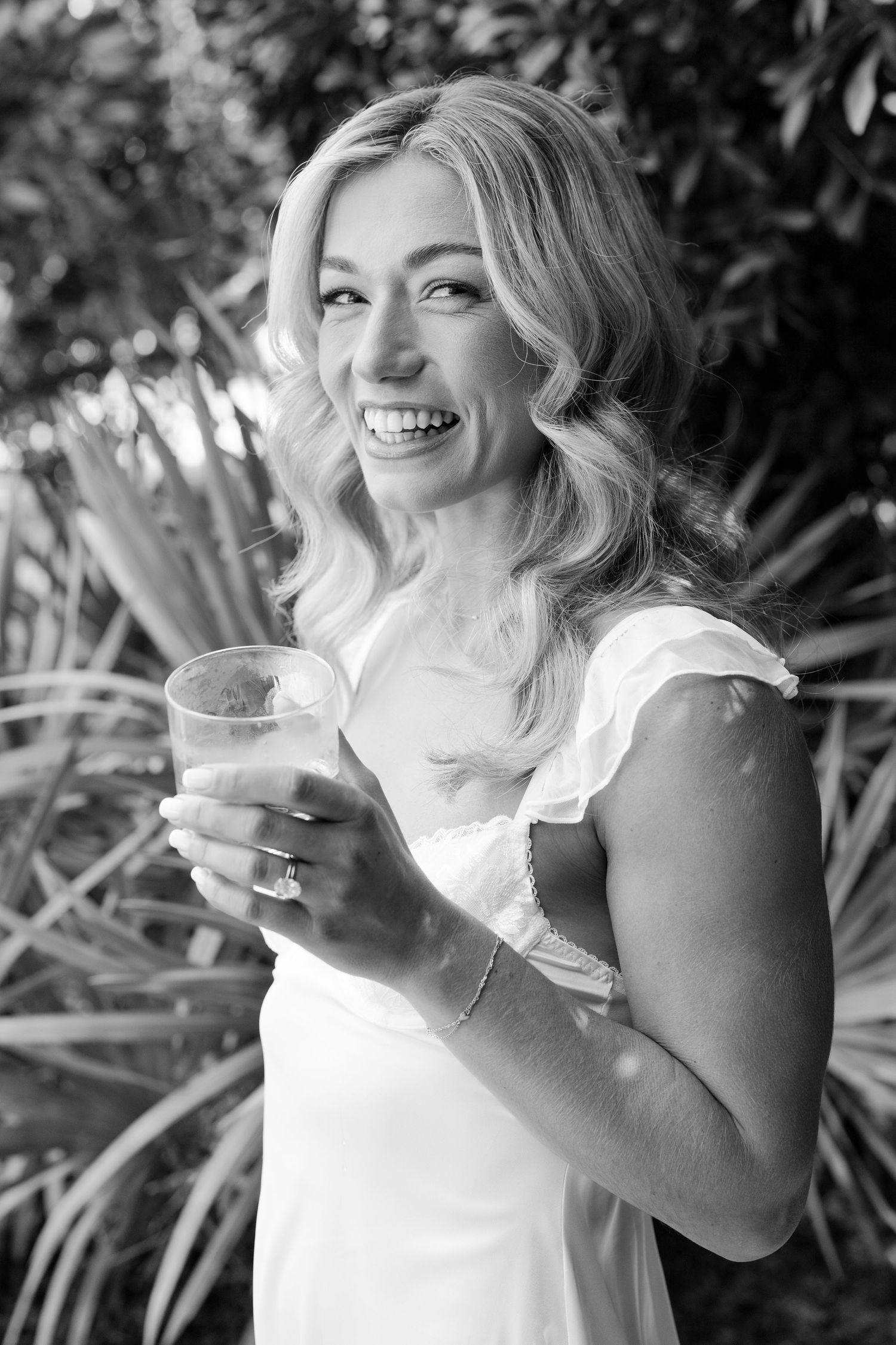 Black and white portrait of a joyful person holding a drink outdoors with natural foliage in background.