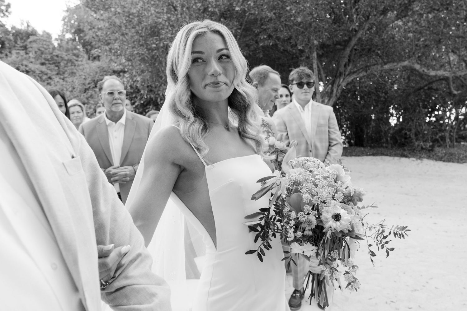A black and white candid photo of wedding party members outdoors with natural greenery in background.