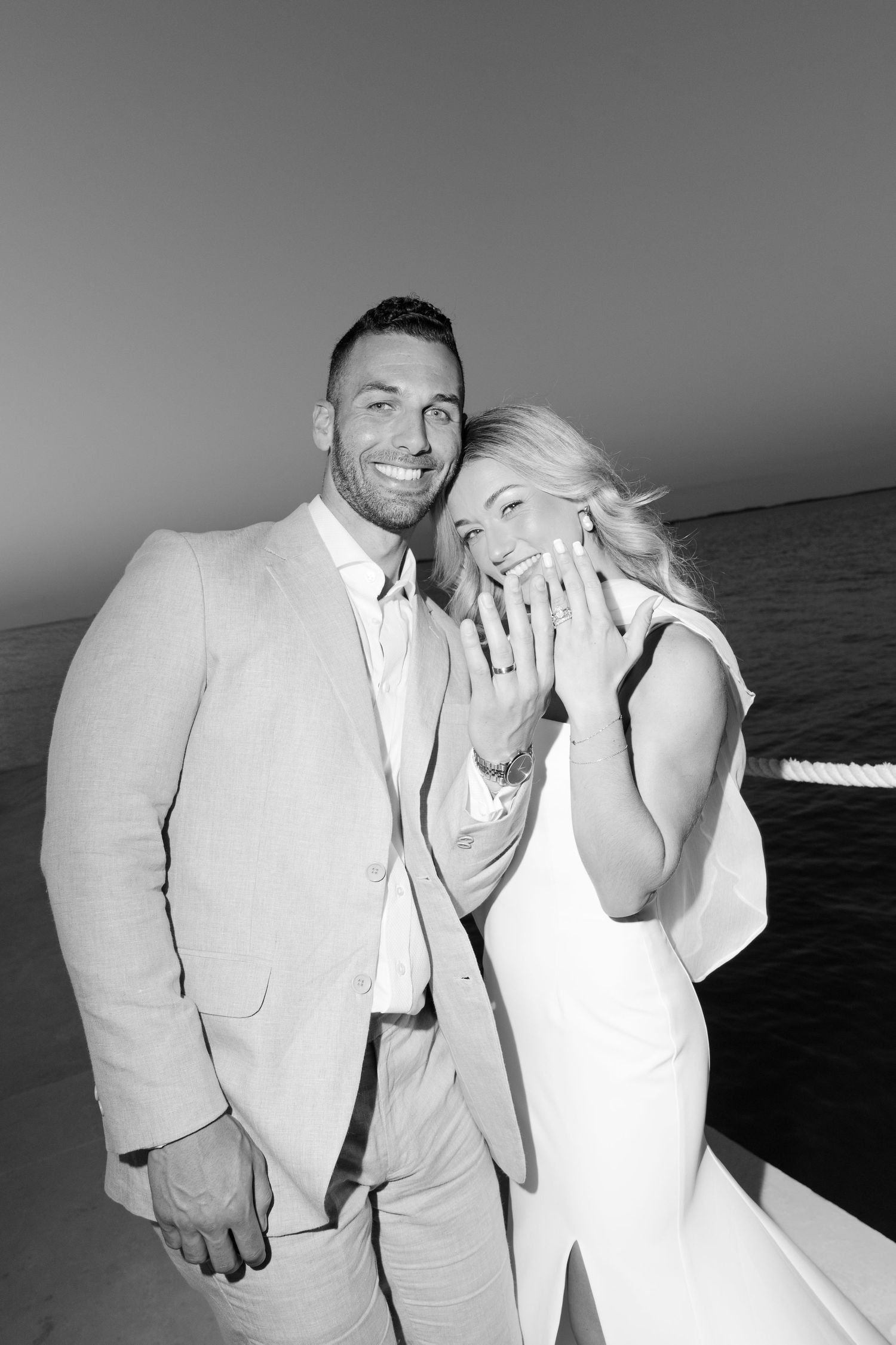 A romantic black and white photo of a couple in formal attire by the ocean at sunset.