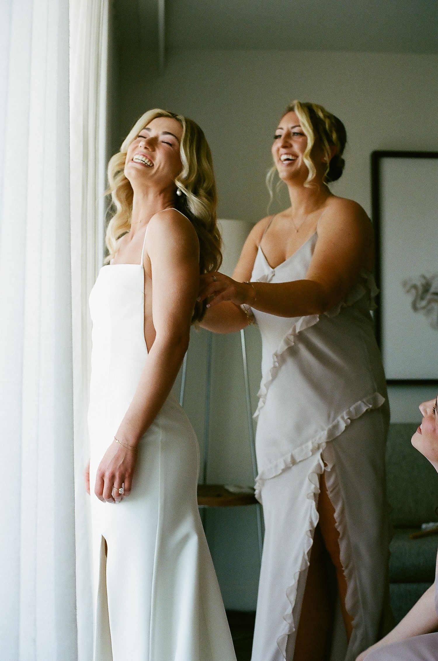 Two women in elegant white dresses share a joyful moment while getting ready by a window.