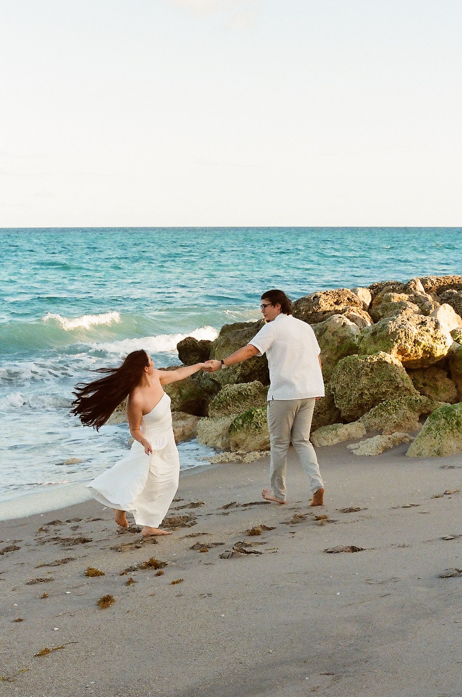 A couple plays on a beach with crashing waves and rocky coastline during a romantic seaside photoshoot.