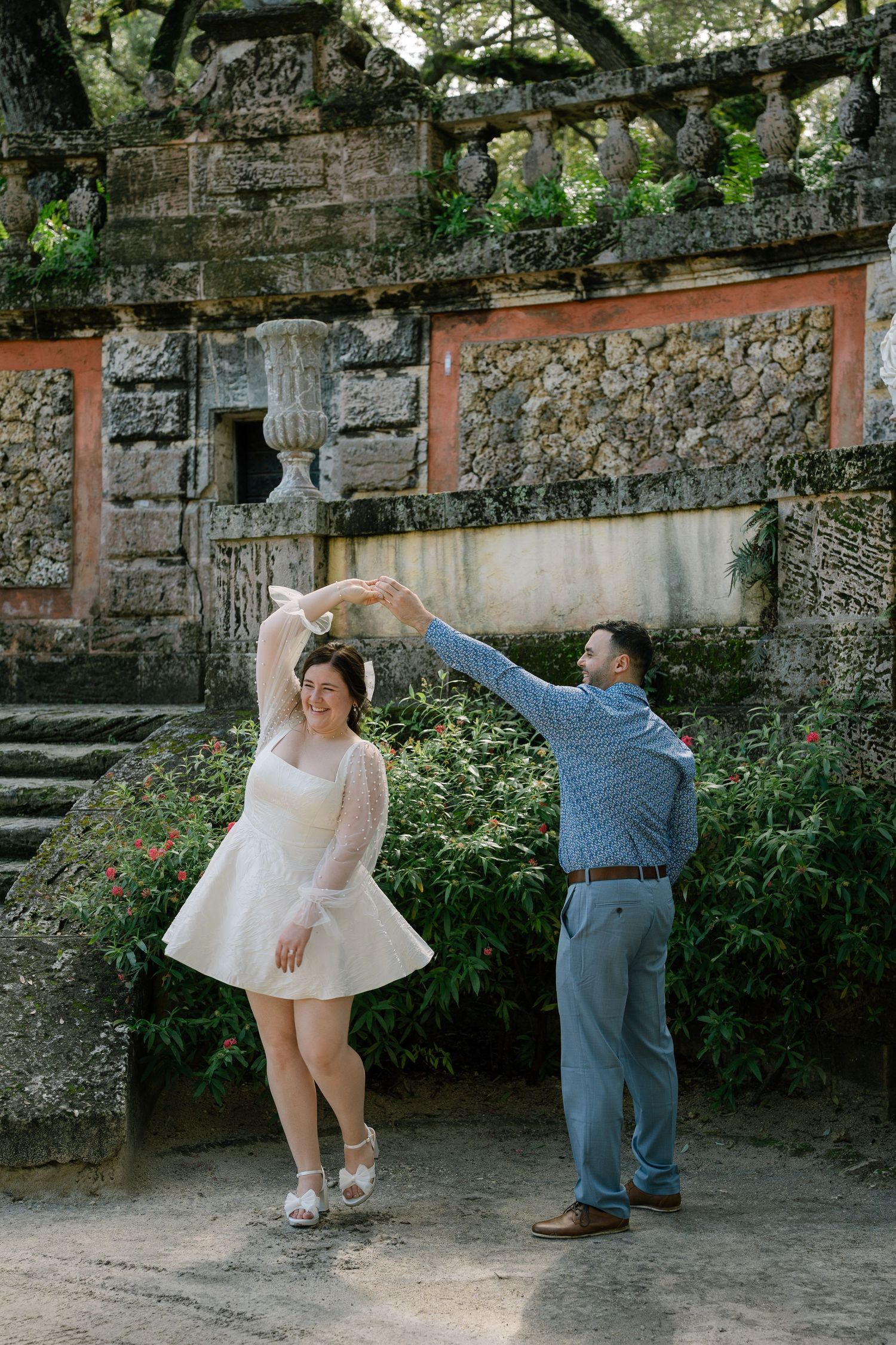 Couple dancing playfully against a rustic stone wall backdrop in a garden setting.