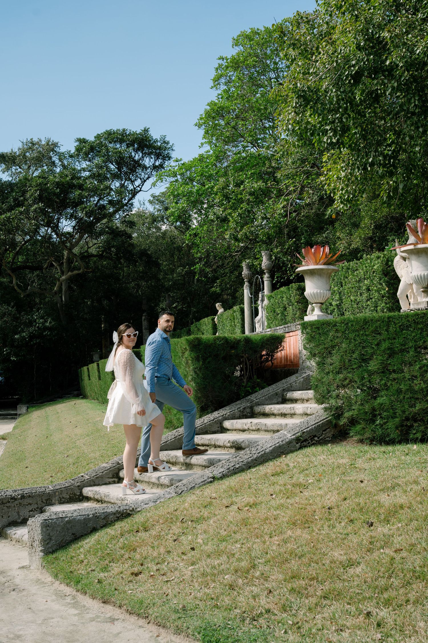 A couple walks up stone garden steps surrounded by manicured hedges and decorative urns.