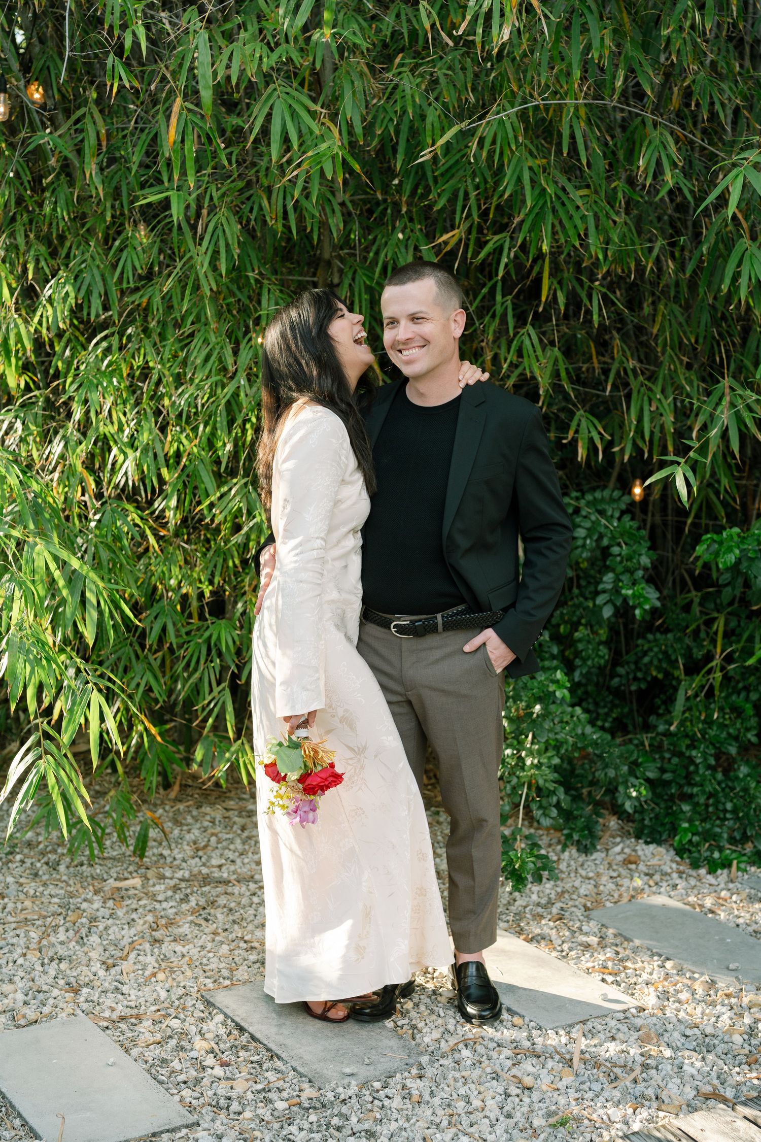 A couple posing together against a bamboo backdrop on a gravel path.