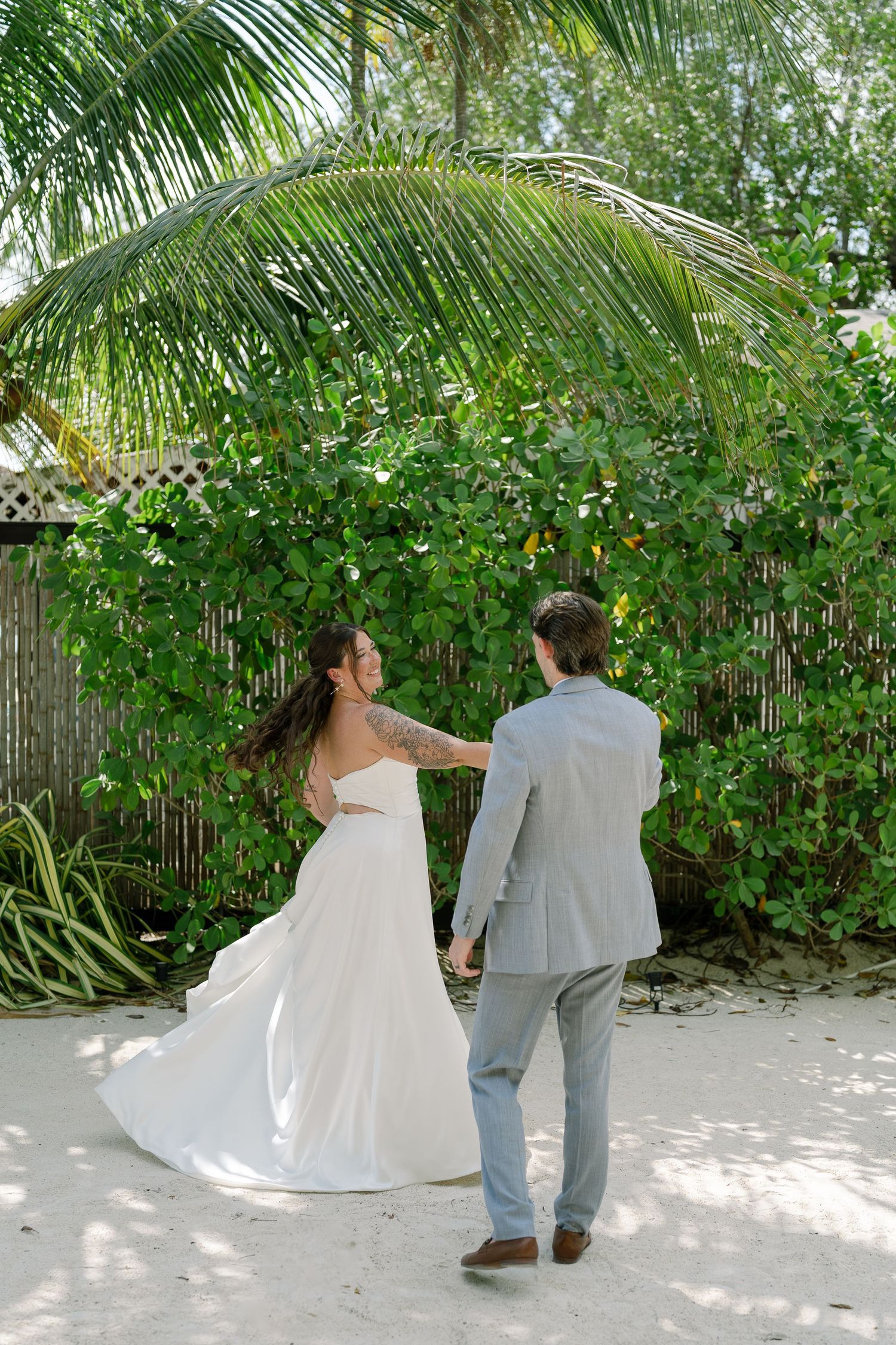 Bride in white gown and groom in gray suit walk hand-in-hand on sandy path beneath tropical palm trees.