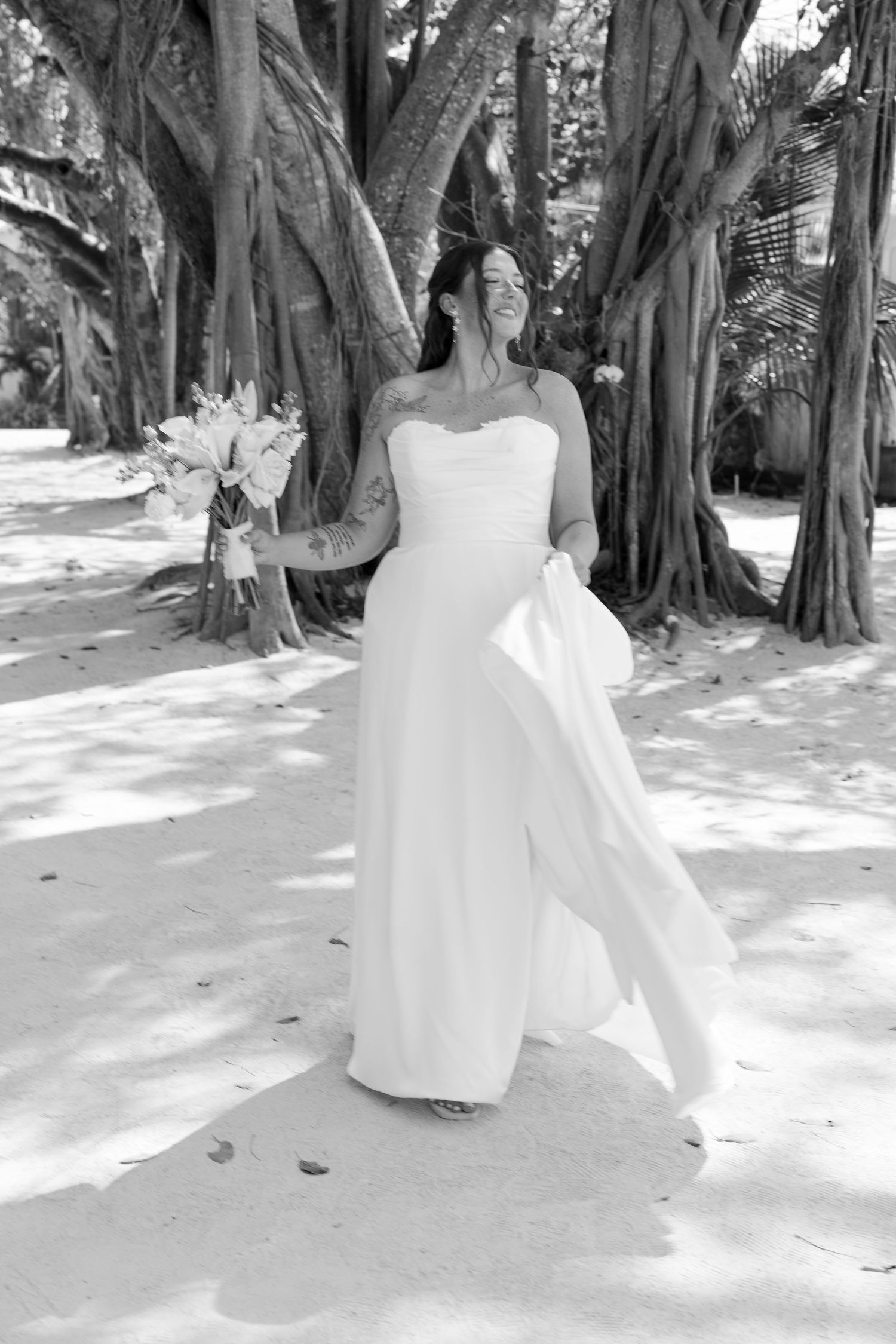 A woman in a white wedding dress stands in a lush, forested area with large trees and greenery surrounding her.