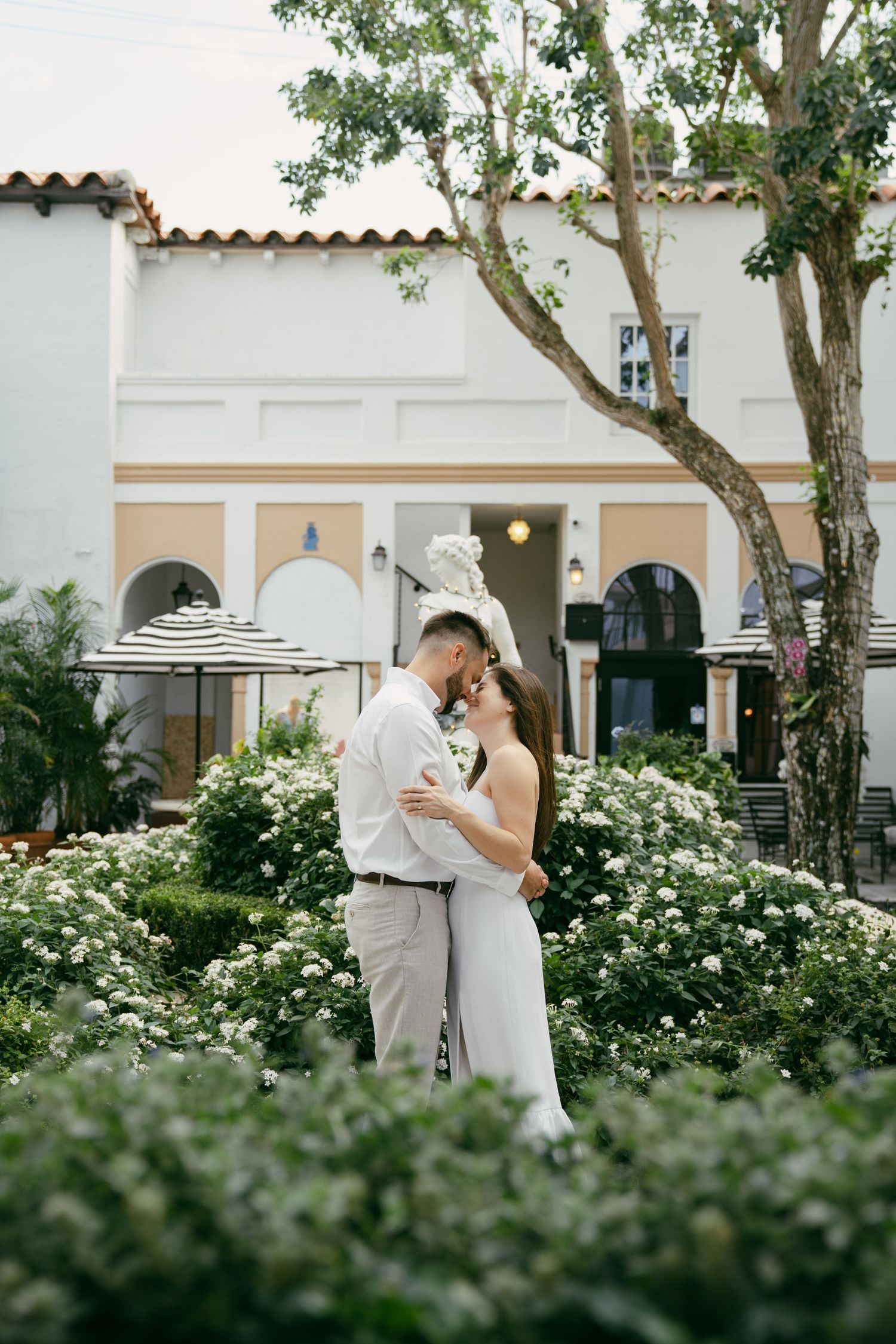 A couple shares a romantic moment in a garden with white roses and Spanish colonial architecture in the background.