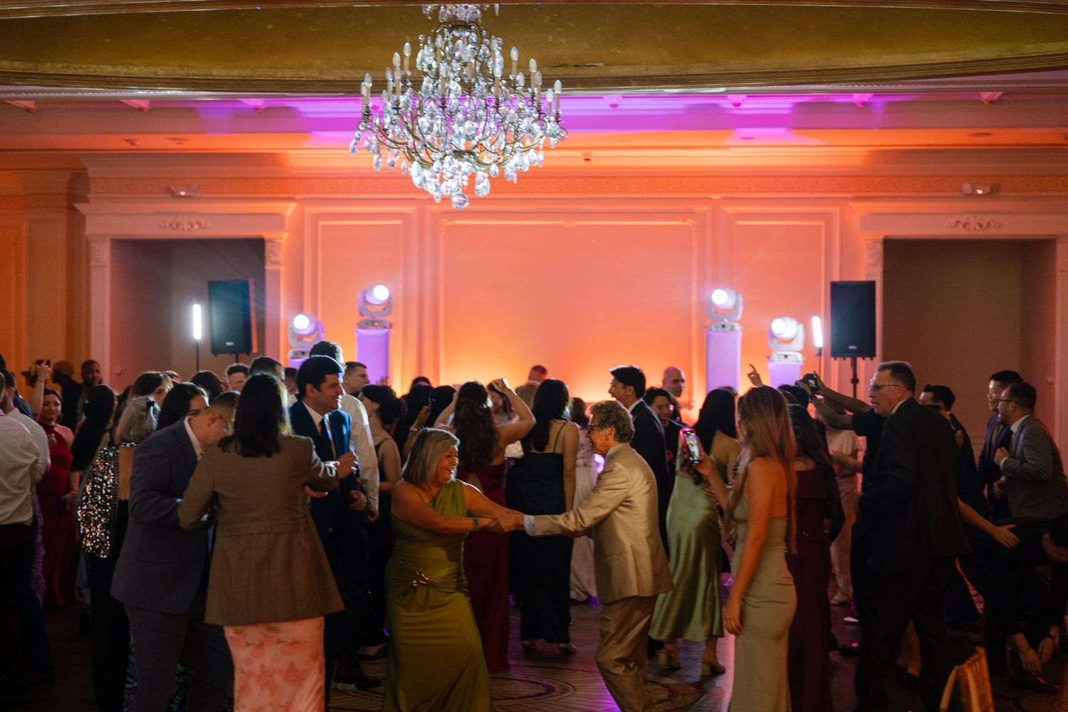 Large crystal chandelier hangs in a ballroom with guests dancing under colorful lighting.