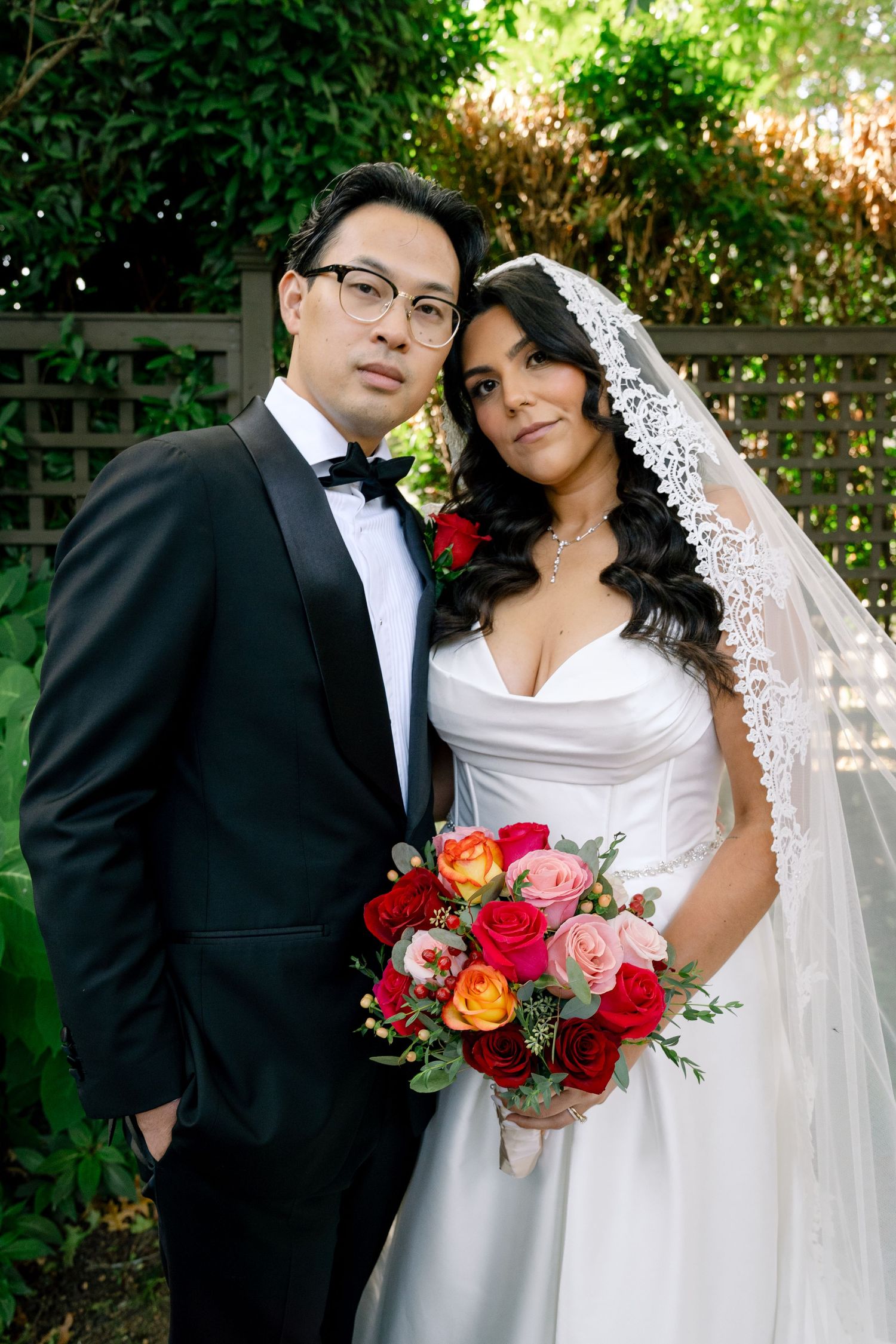 A bride in a white dress holds a vibrant red and pink bouquet while posing with her groom in a black tuxedo outdoors.