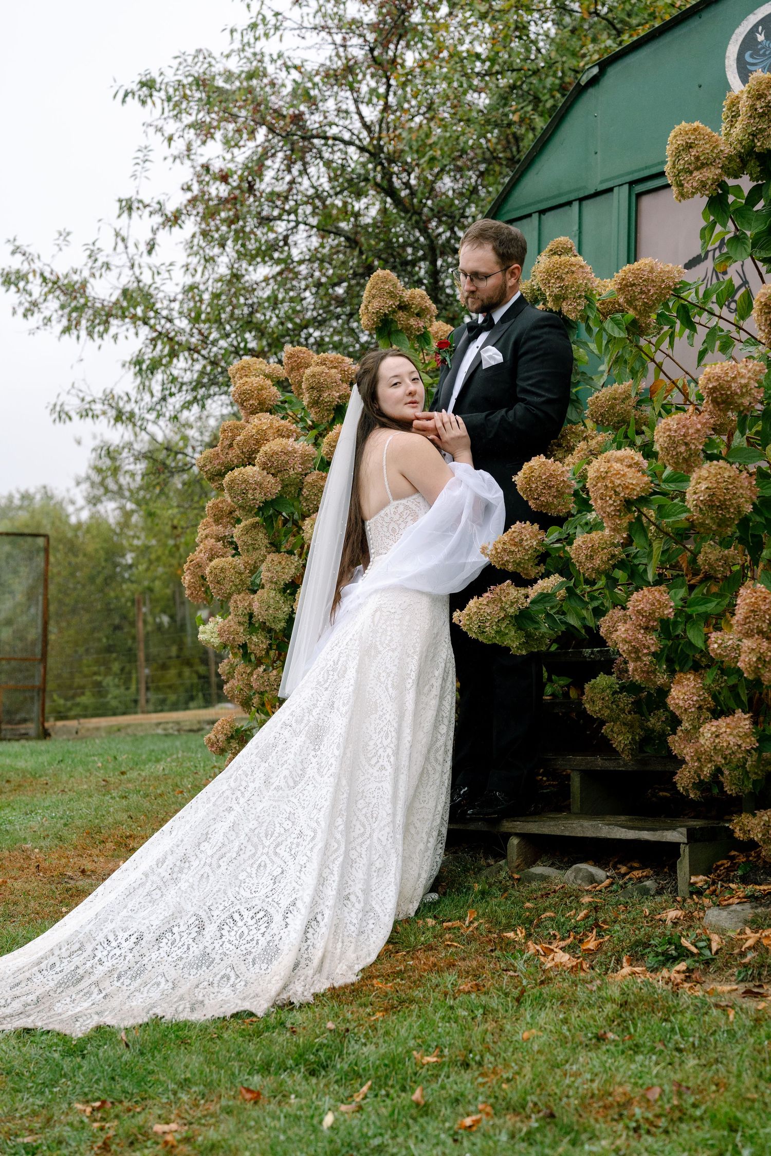 Romantic wedding couple poses together against green wall with dried hydrangea flowers in garden setting.