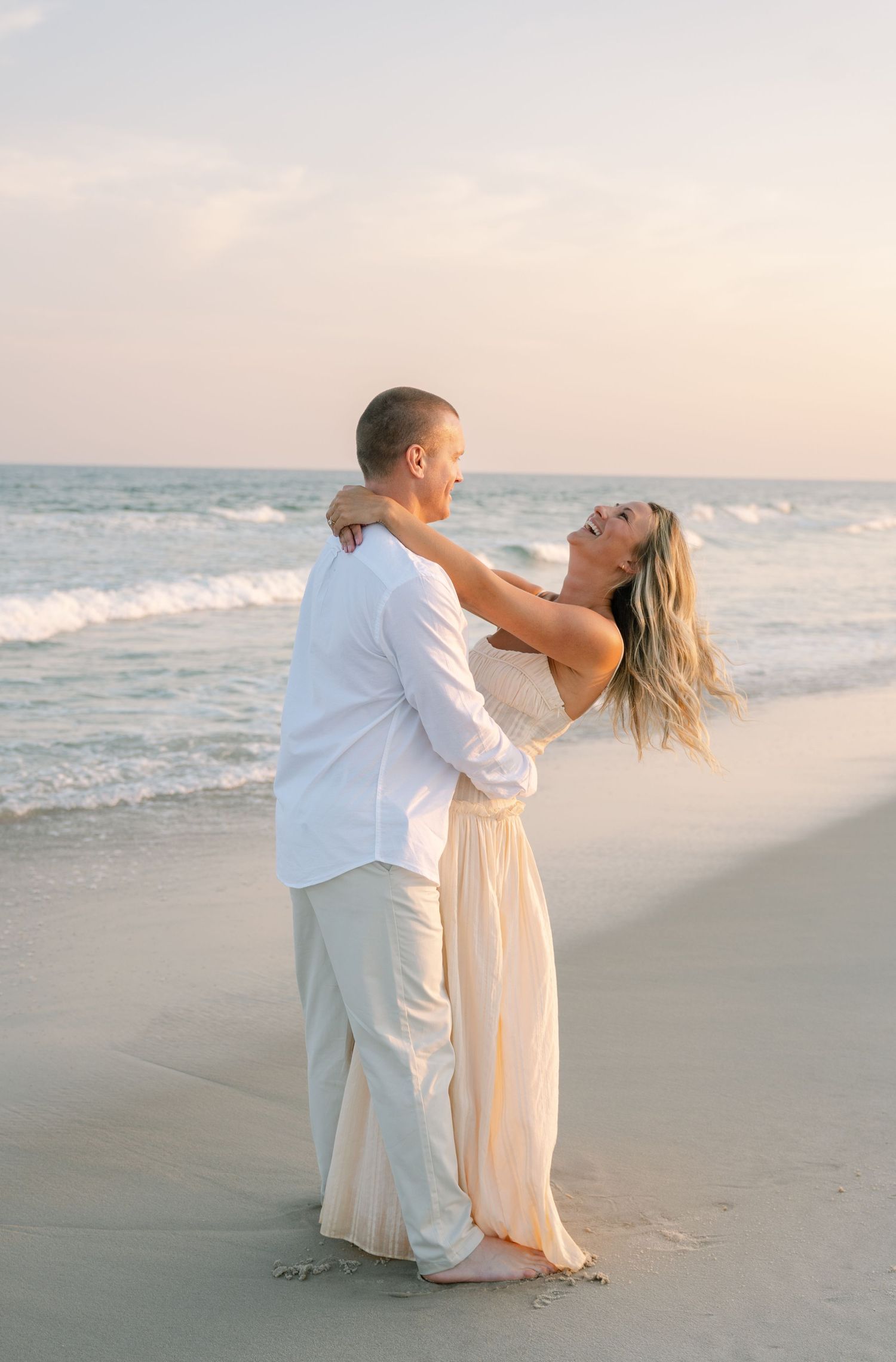 A couple shares a romantic moment together on a sandy beach at sunset, dressed in white and cream colored outfits.