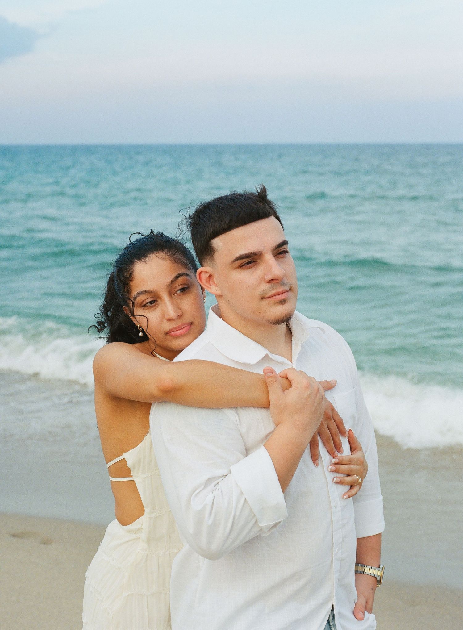 A couple embraces on a beach with turquoise waters and gentle waves in the background.