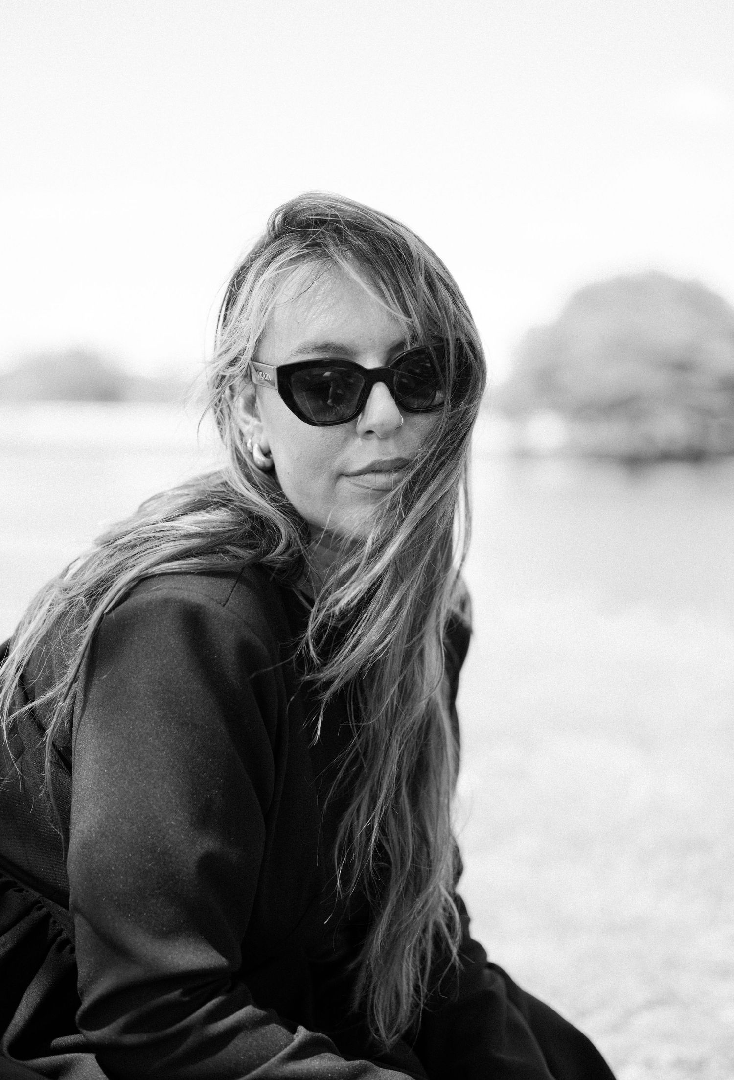 A black and white portrait of someone wearing sunglasses and a dark sweater with windswept hair outdoors.