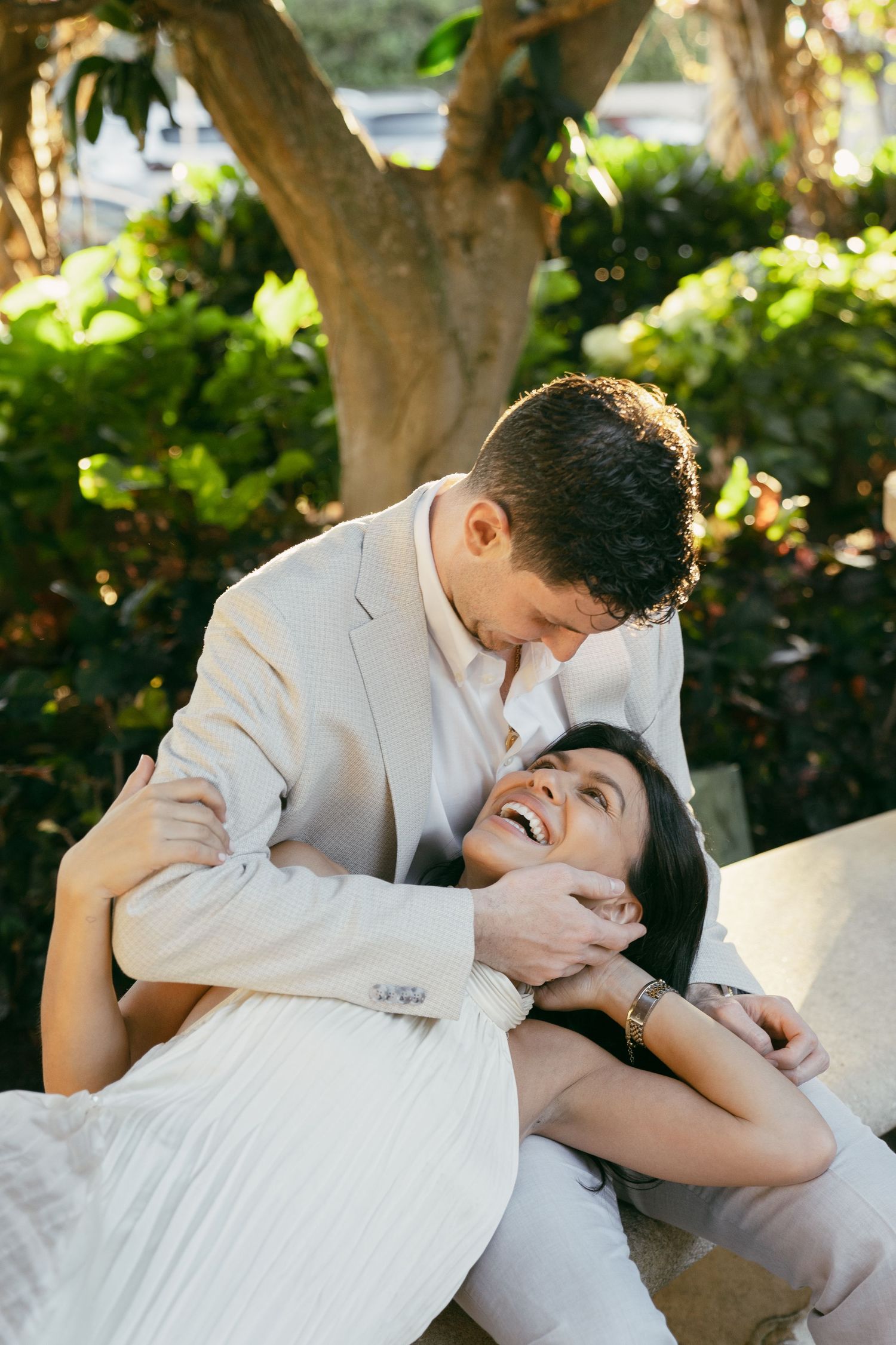 A couple in white clothing embrace intimately while lounging outdoors in a garden setting with dappled sunlight.