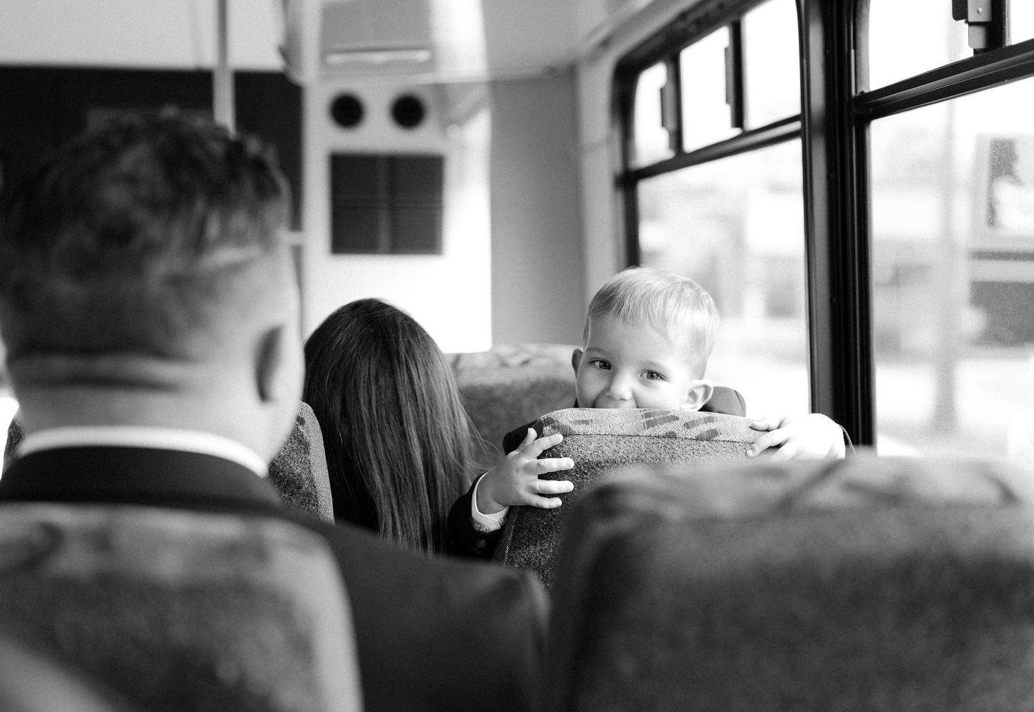 A small child peers over the back of a bus seat in black and white while other passengers sit facing forward.