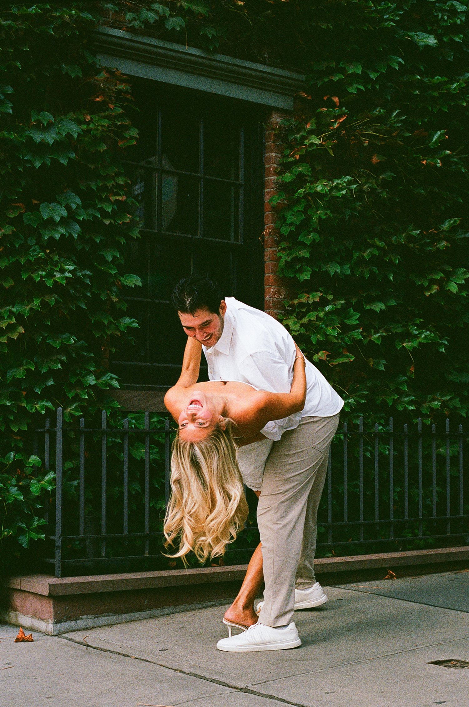 Couple sharing a romantic dip pose on a city sidewalk, surrounded by lush green ivy-covered brick wall.