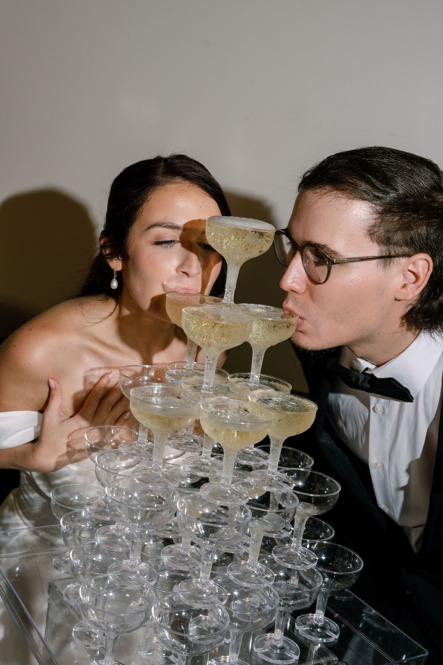 Bride and groom sipping champagne from a tiered tower of coupe glasses at their wedding reception.