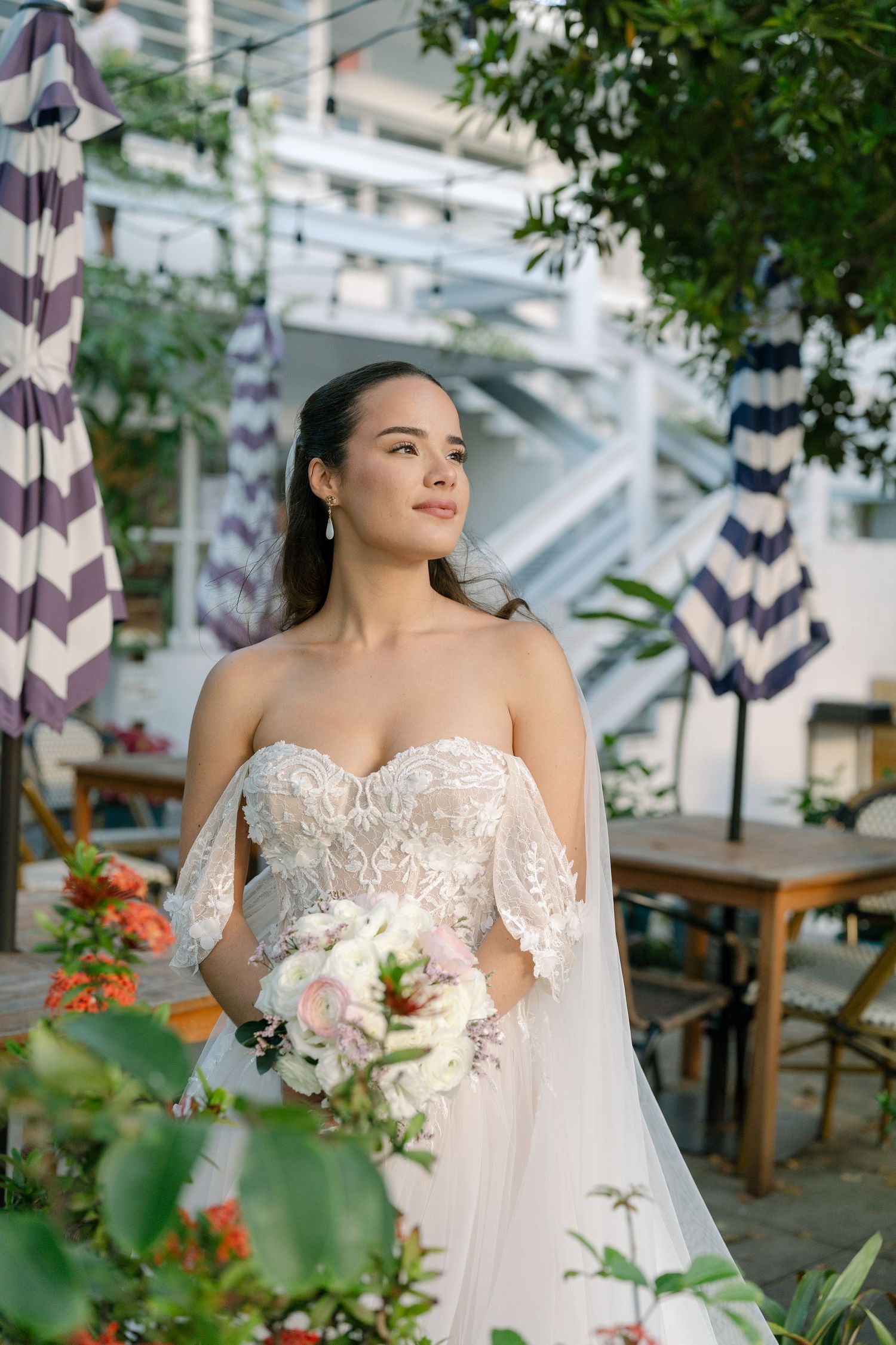 Elegant bride in strapless lace wedding gown holding floral bouquet outdoors among lush greenery.