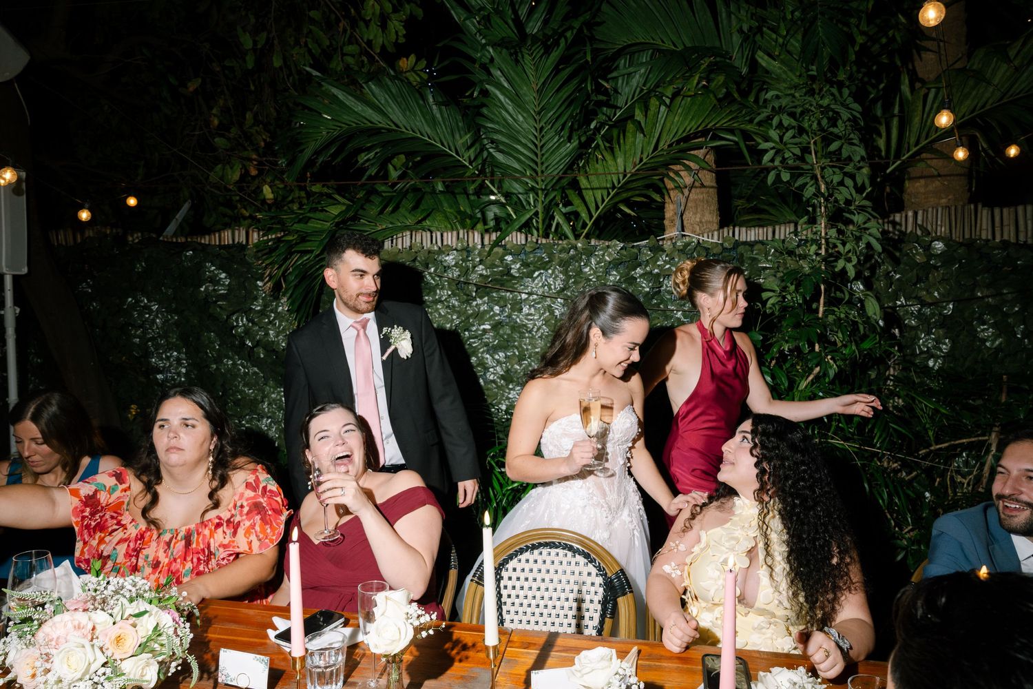 Bride and groom celebrate with wedding party guests at decorated reception table surrounded by tropical greenery.
