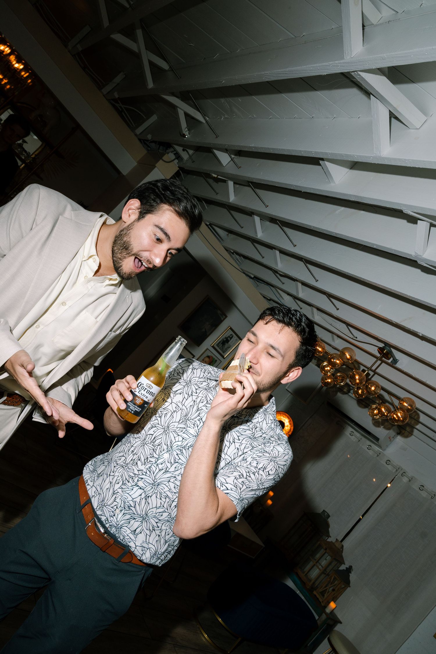 Two men laugh together at wedding reception, one holding a beer bottle indoors under wooden ceiling.
