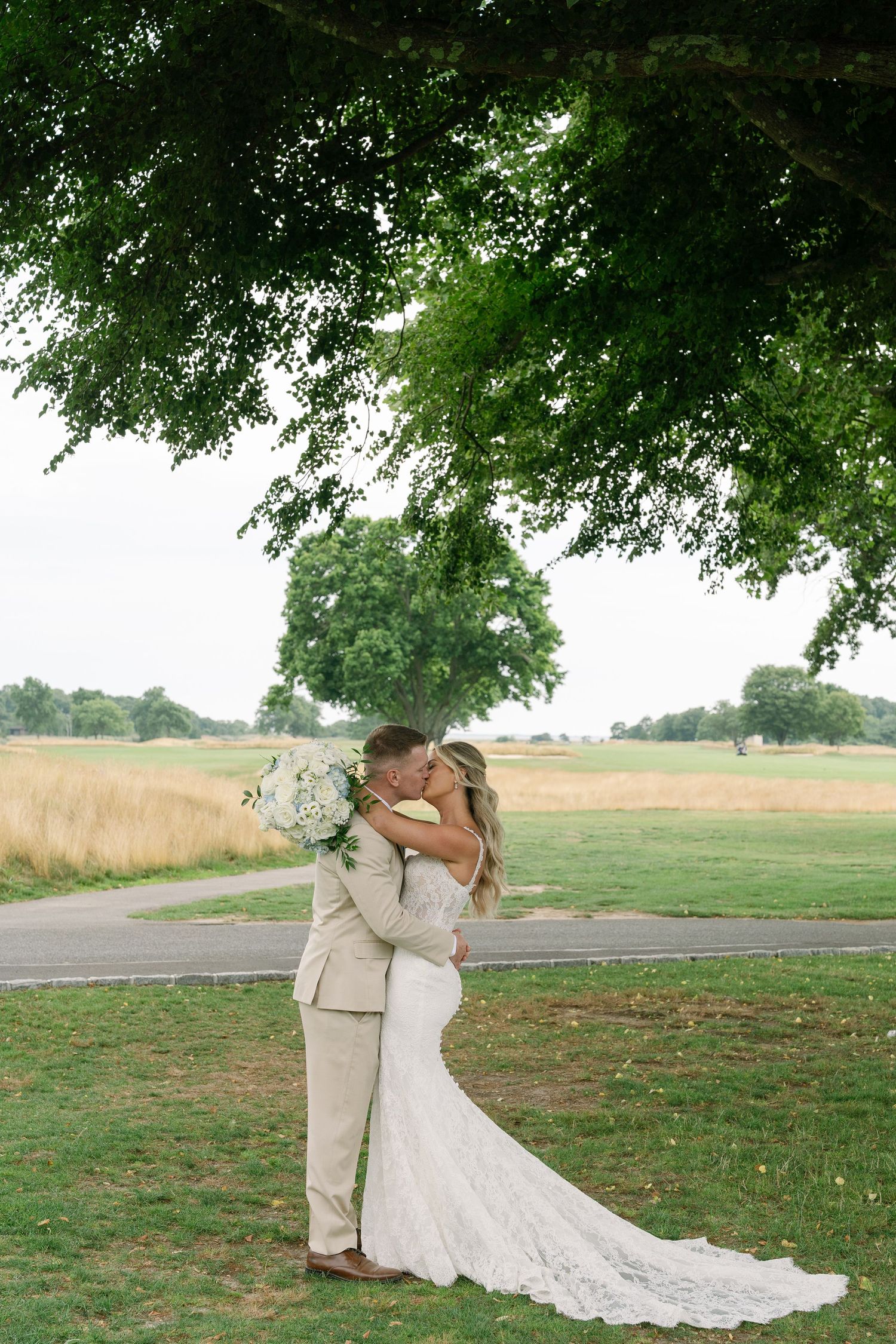 Newlywed couple shares a romantic kiss under a lush green tree on an open grassy field on their wedding day.