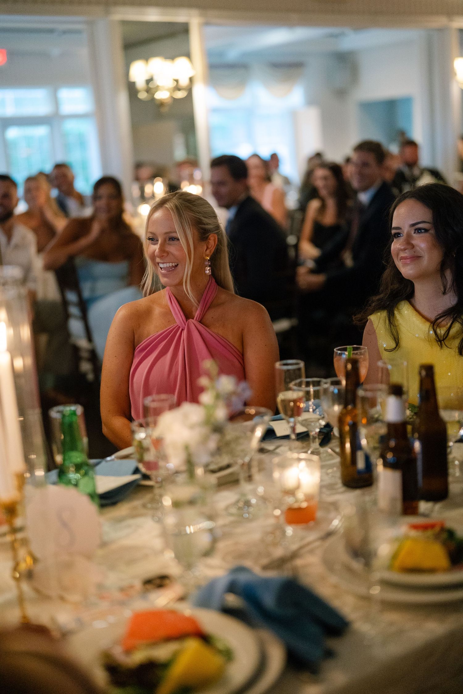 A smiling woman in a pink halter dress sits at an elegantly set wedding reception dinner table.