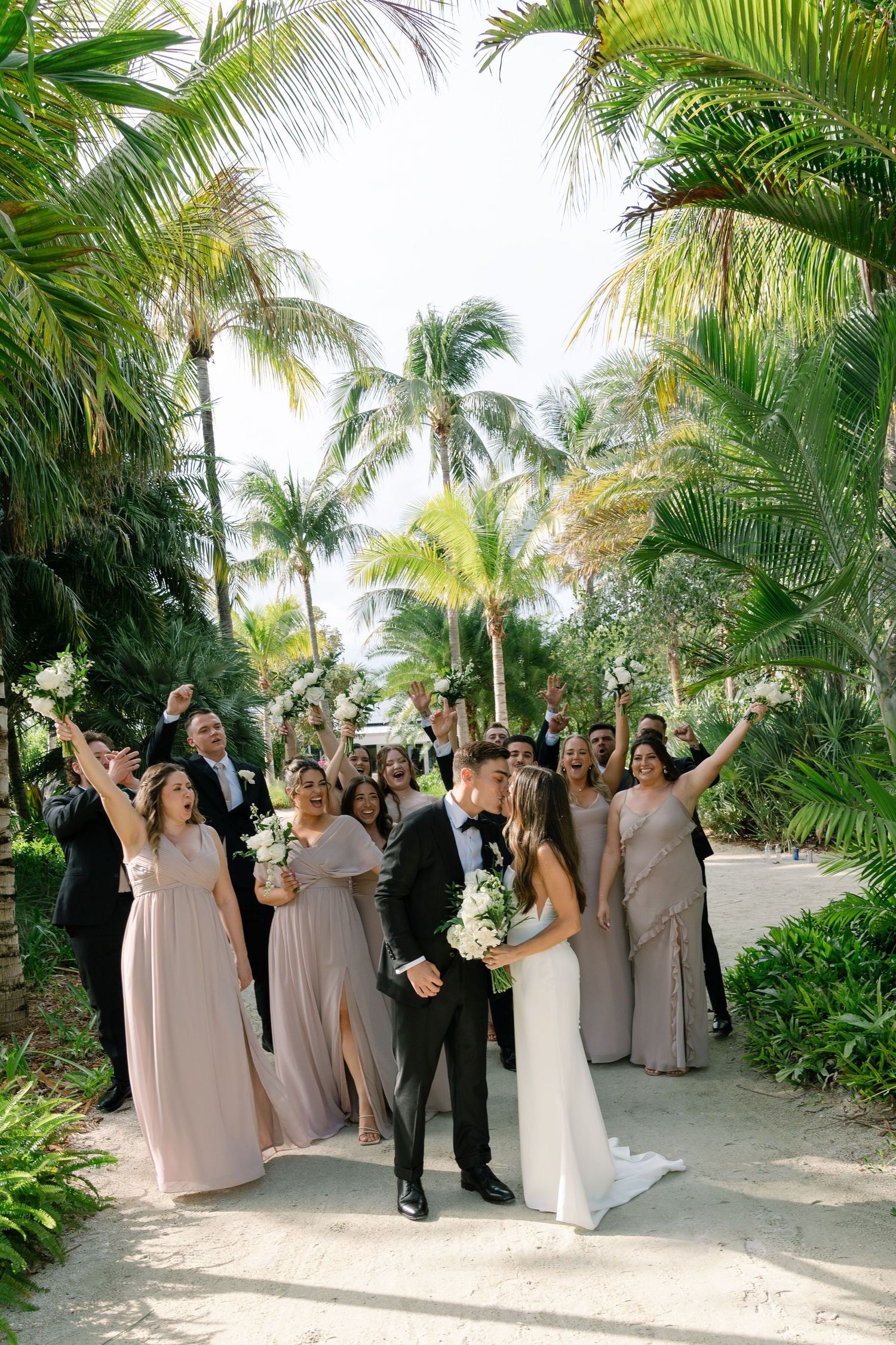 Joyful wedding party celebrating with the bride and groom on a sandy path surrounded by tropical palm trees.