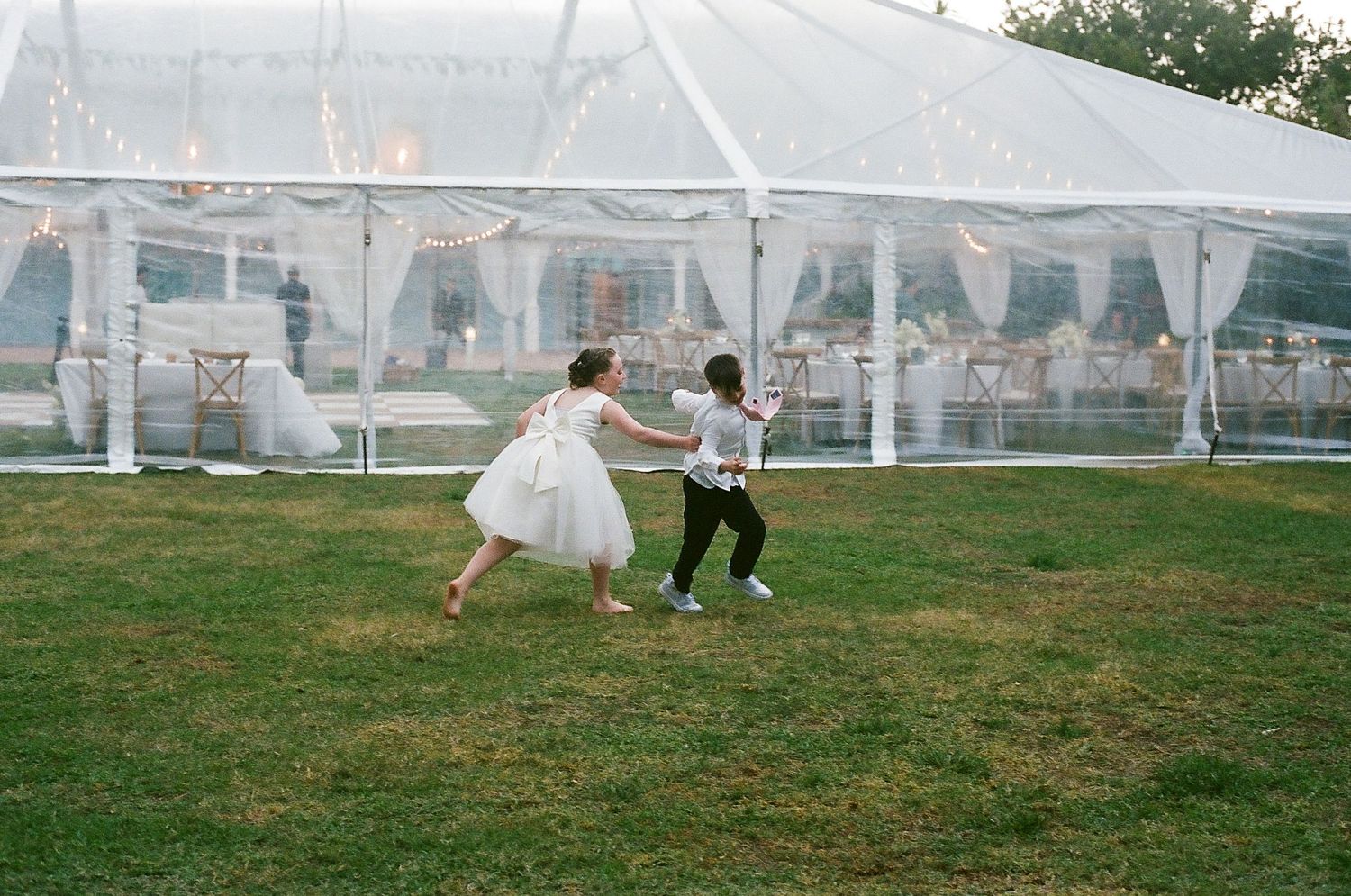 Two children in wedding attire dance playfully on green grass outside a white tent reception venue.