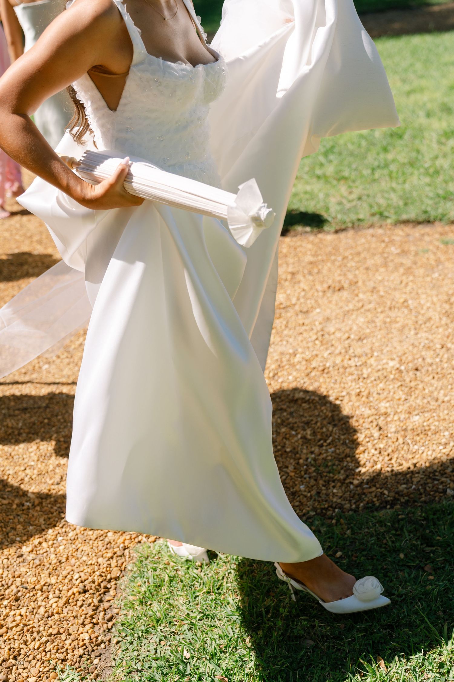Bride in white satin gown and heels holding a folded fan while walking on a gravel path at an outdoor wedding.