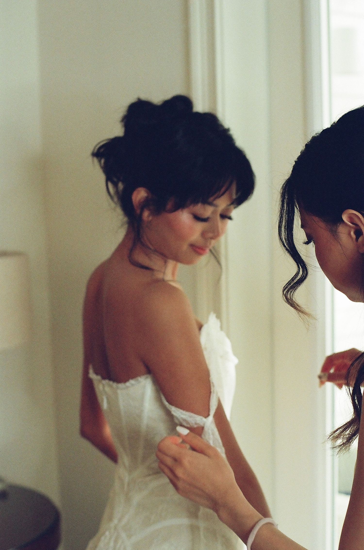 A bride in a white strapless gown having her dress fastened by a bridesmaid near a bright window.